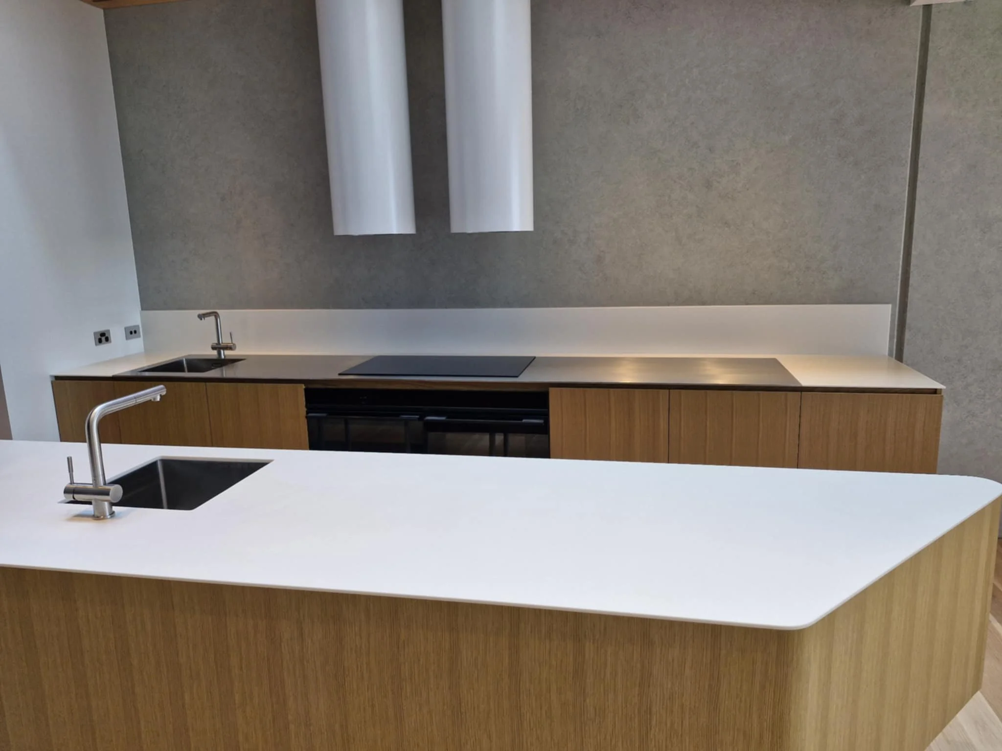 Modern white kitchen with minimalist design, featuring an island with a double sink, backsplash with herringbone pattern, and upper cabinets. Kitchen appliances include a coffee maker and a stove, with a small nook containing a sink visible on the right.