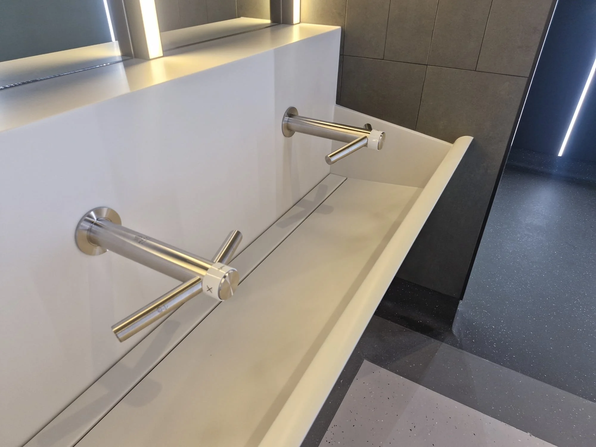 A white changing table with metal safety rails on both sides, located in a modern facility with dark tiled walls and a speckled black and gray floor.