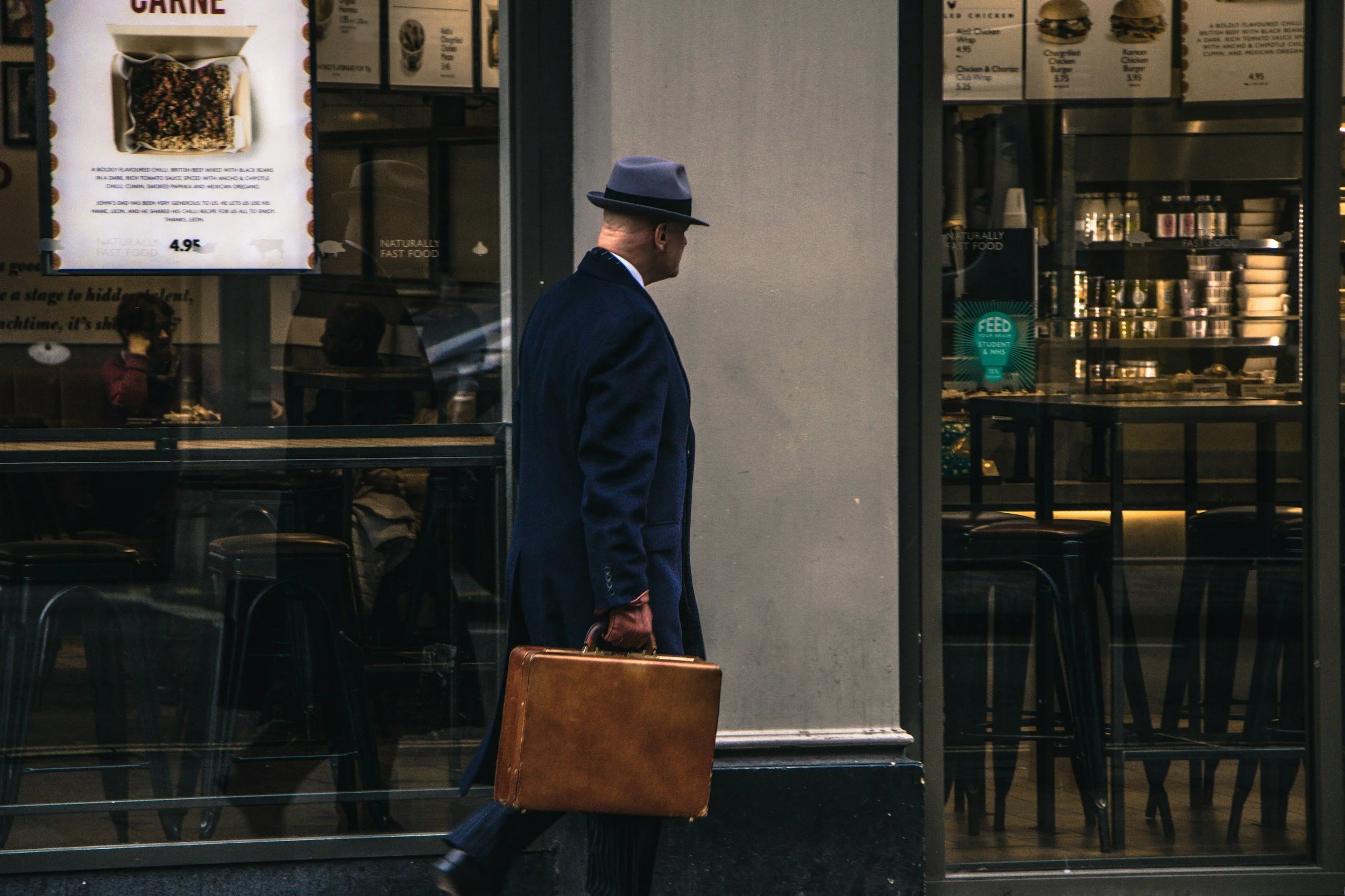A man in a dark suit and gray hat walking past a glass storefront, carrying a brown leather briefcase.