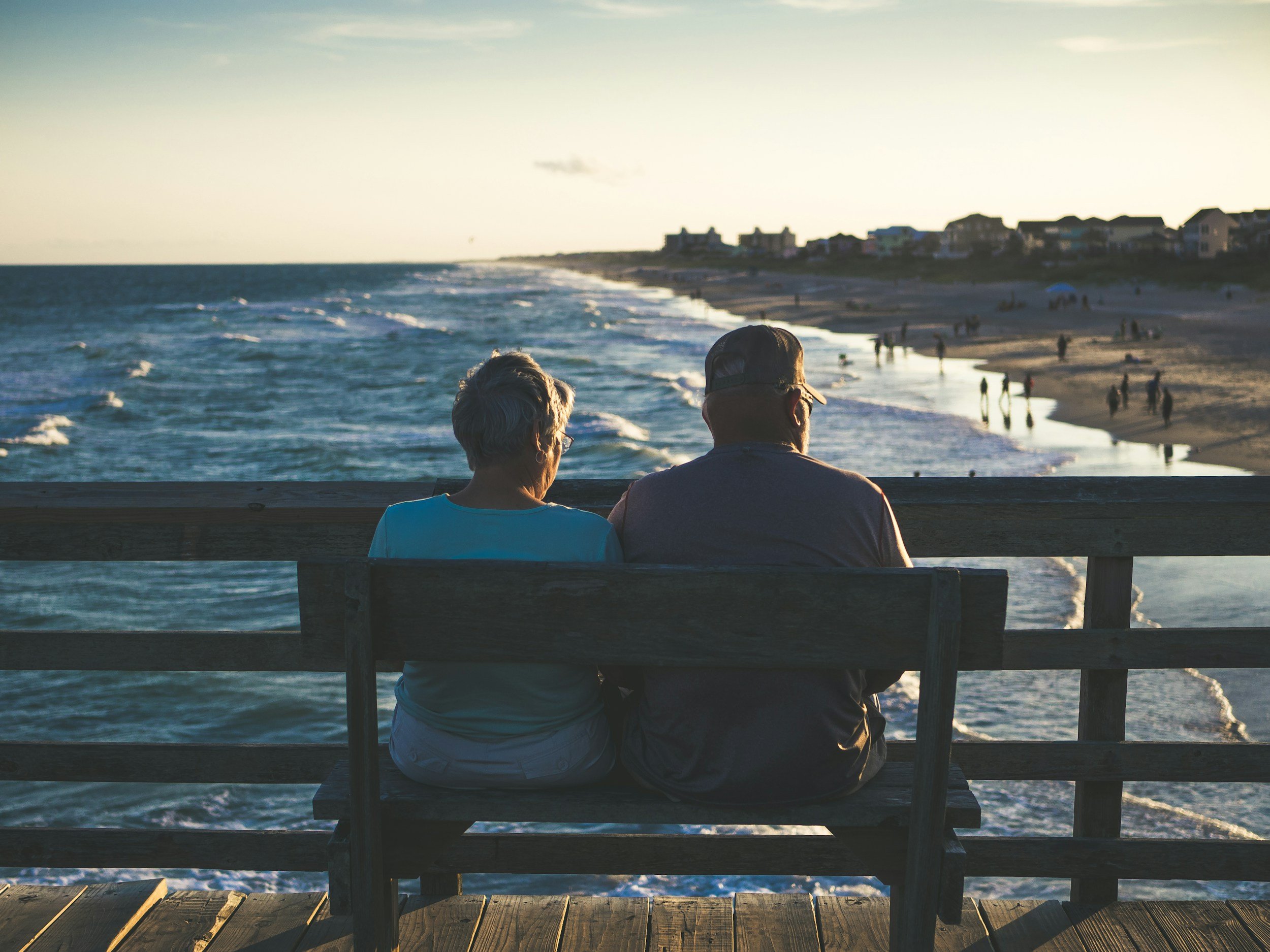An elderly couple sitting on a wooden bench, facing the ocean, watching the sunset at a beach pier.