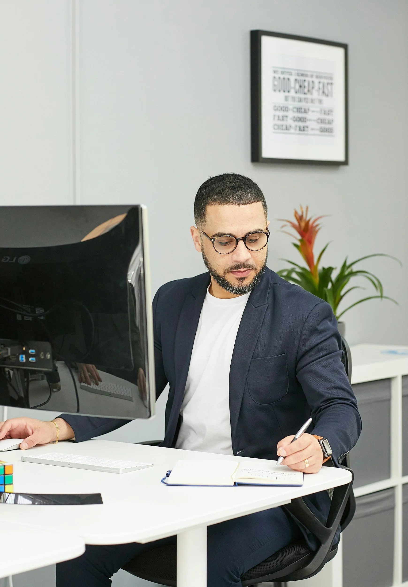 Man in a navy blazer and white shirt sitting at a white desk, working with a notebook, pen, computer monitor, and a mini Rubik's Cube in an office with gray walls and a potted plant.