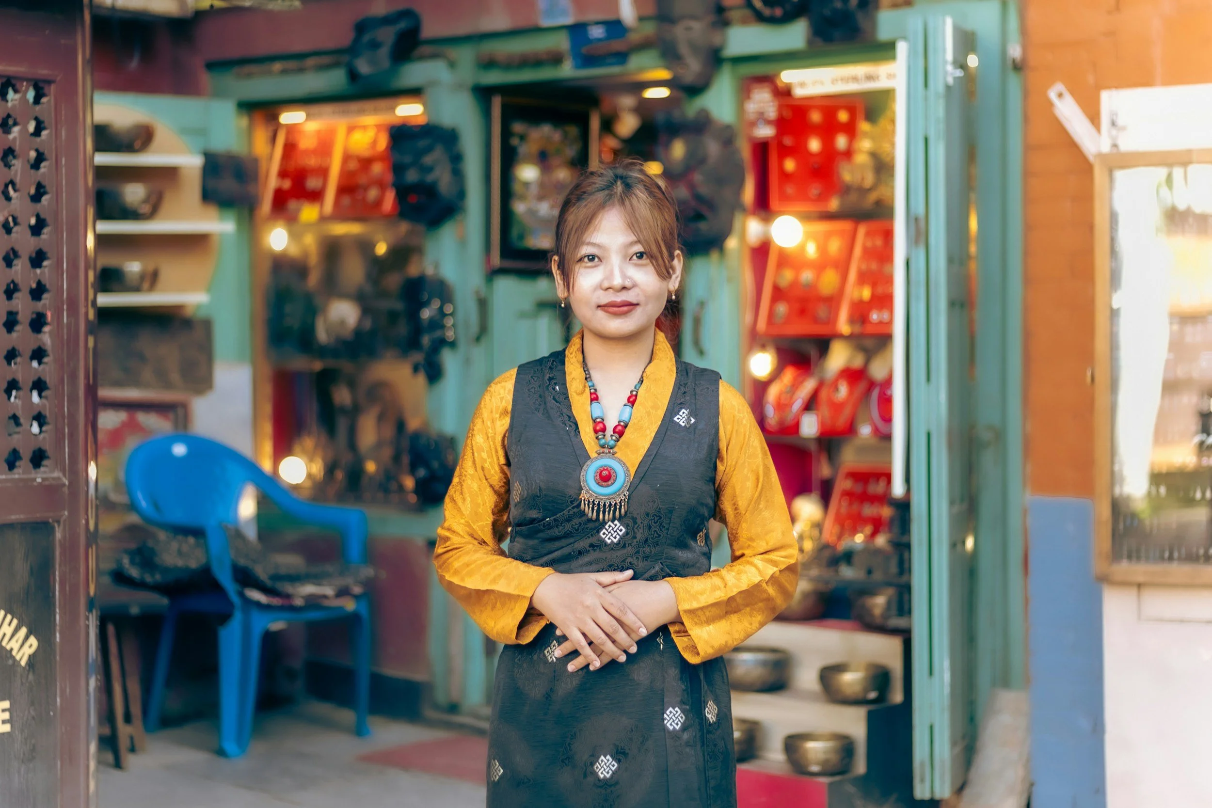 A woman in a traditional black and yellow outfit stands in front of a shop filled with colorful items and jewelry, smiling at the camera.