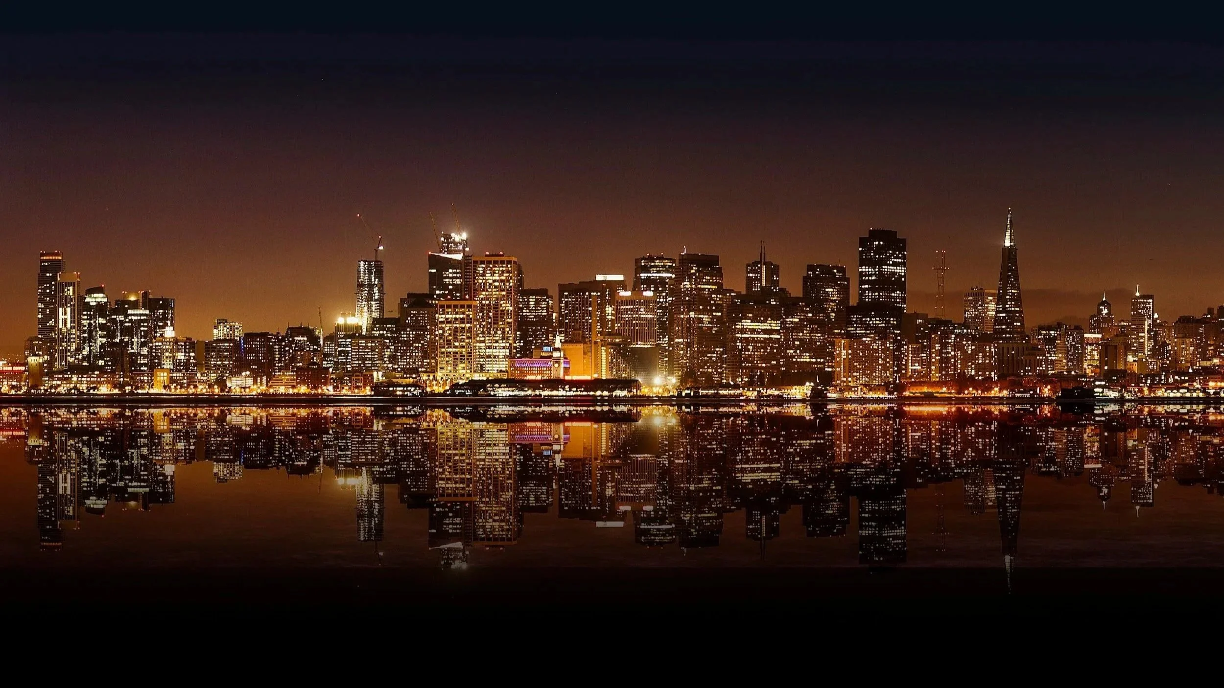 Nighttime city skyline of San Francisco with illuminated buildings and their reflection in the water.