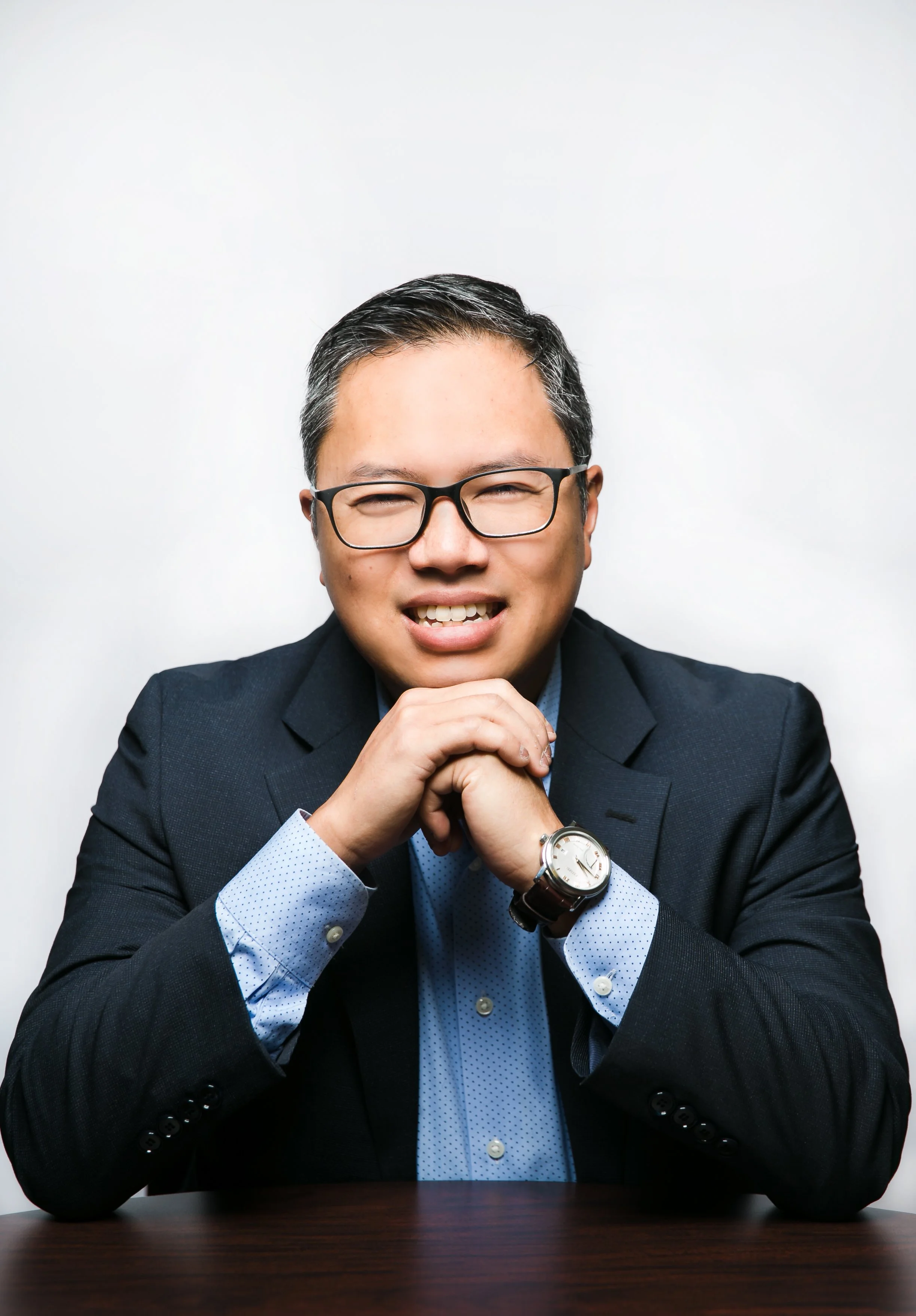 A man in a suit and glasses sitting at a table, smiling with hands clasped, wearing a watch, against a plain white background.