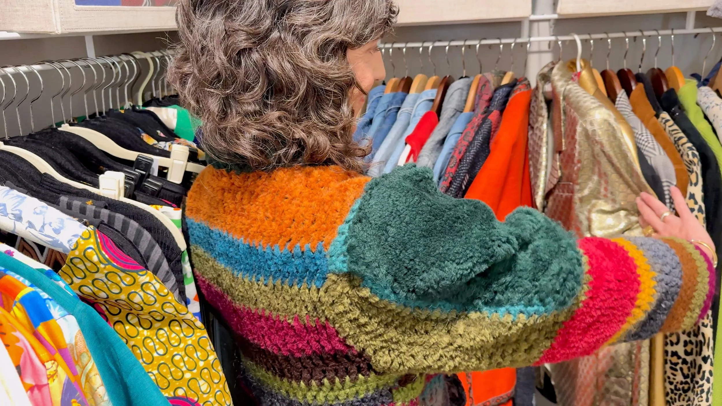 Rajka, a woman with curly hair wearing a multicolored fuzzy sweater shopping for clothes, looking at garments on hangers in her closet.