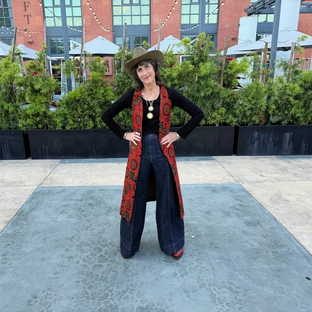 Rajka in wide-leg jeans and a black shirt stands outdoors wears ing wide-brimmed hat, layered necklaces, and a long, red patterned vest. She has dark curly hair and is posing with her hands on her hips.