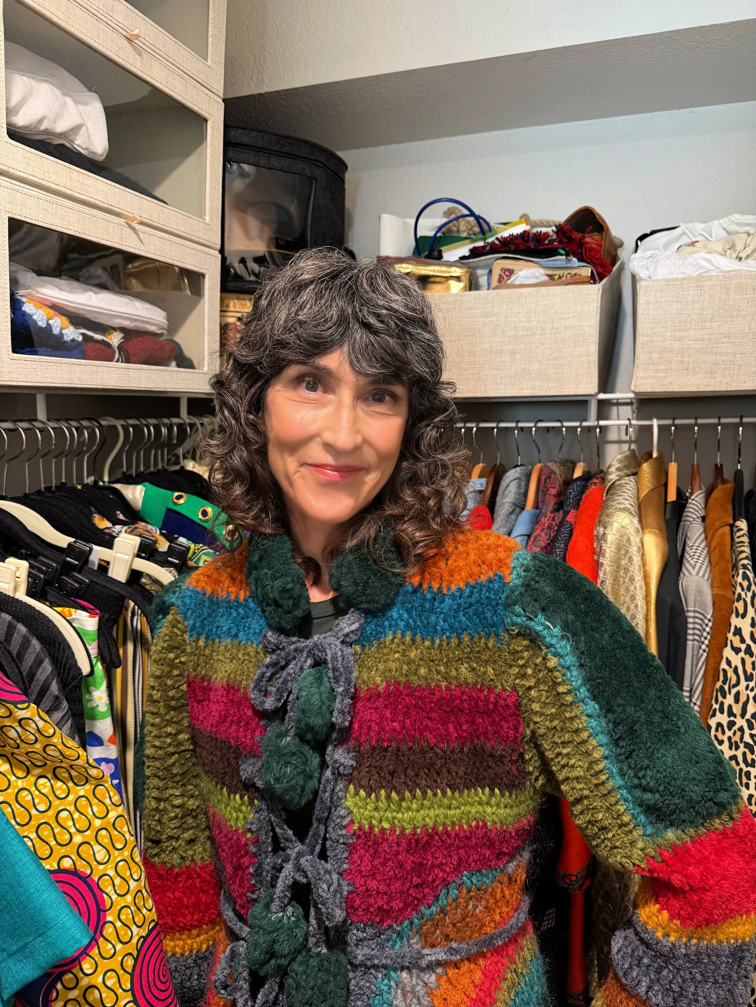 Rajka, a woman with curly dark hair wearing a multicolored, striped, fuzzy sweater standing in a closet with clothes, storage bins, and boxes behind her.