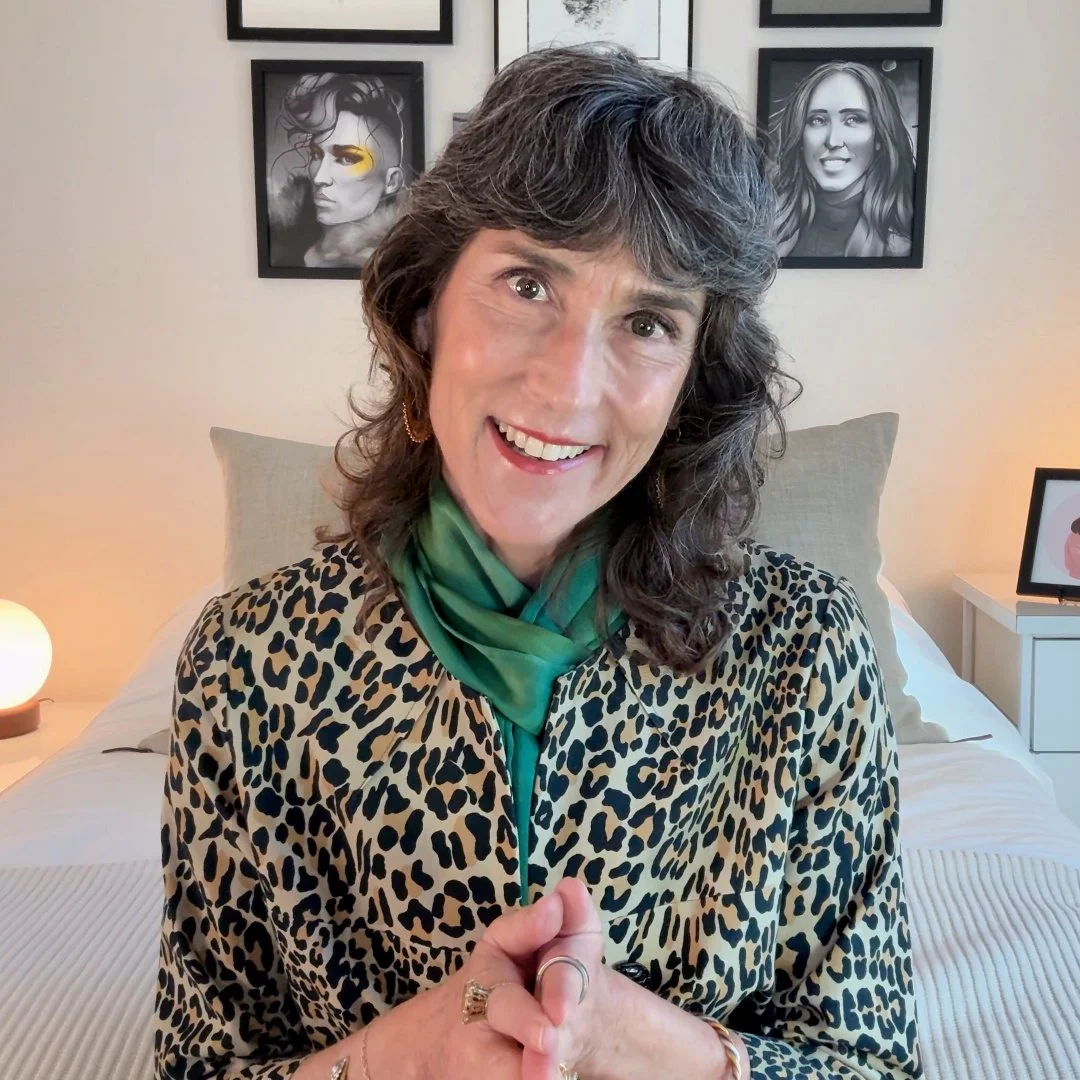 Rajka, a woman with curly gray hair smiling while sitting in a bedroom, wearing a leopard print top and a green scarf, with black and white framed photos on the wall behind her.