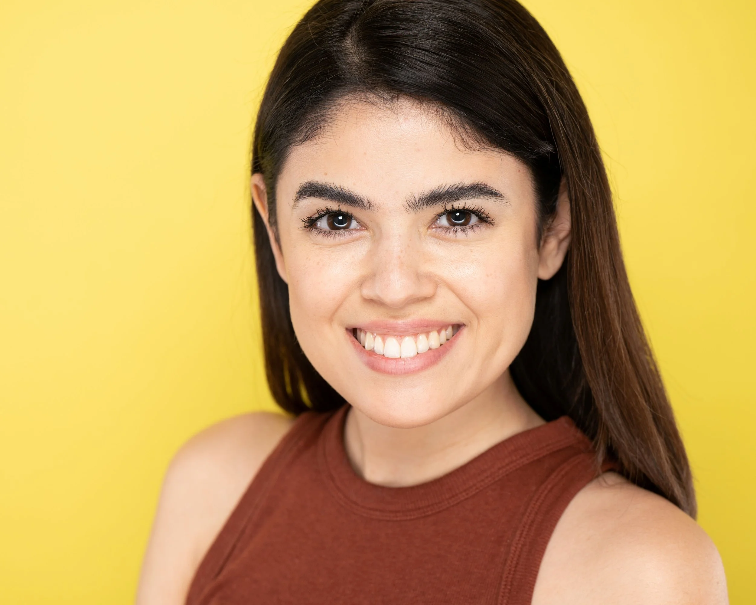 A young woman actress with dark brown hair smiling, wearing a sleeveless rusty red top, against a yellow background.