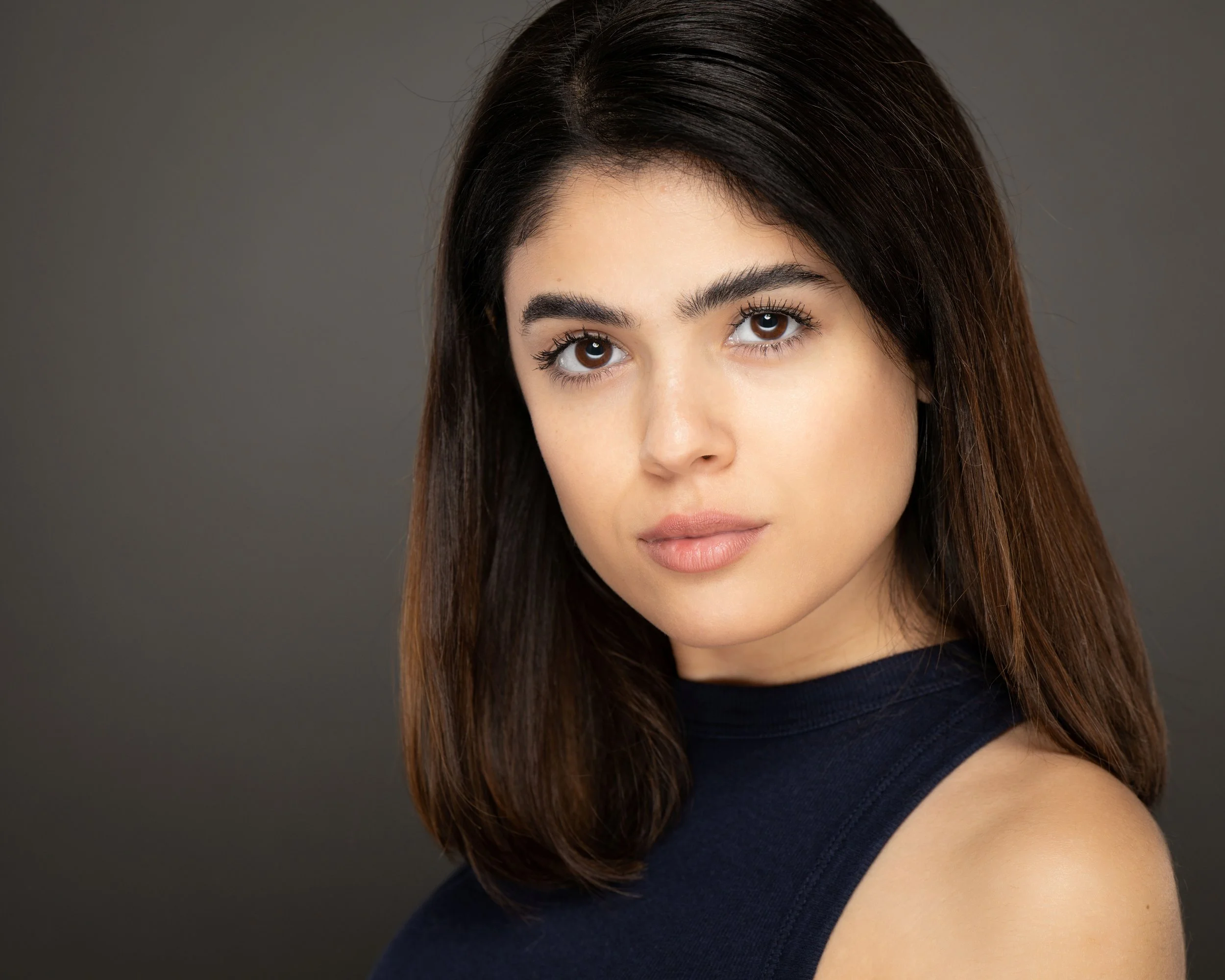 Close-up of a young woman actress with dark brown hair, brown eyes, and light skin, wearing a sleeveless black top, posing against a plain gray background.
