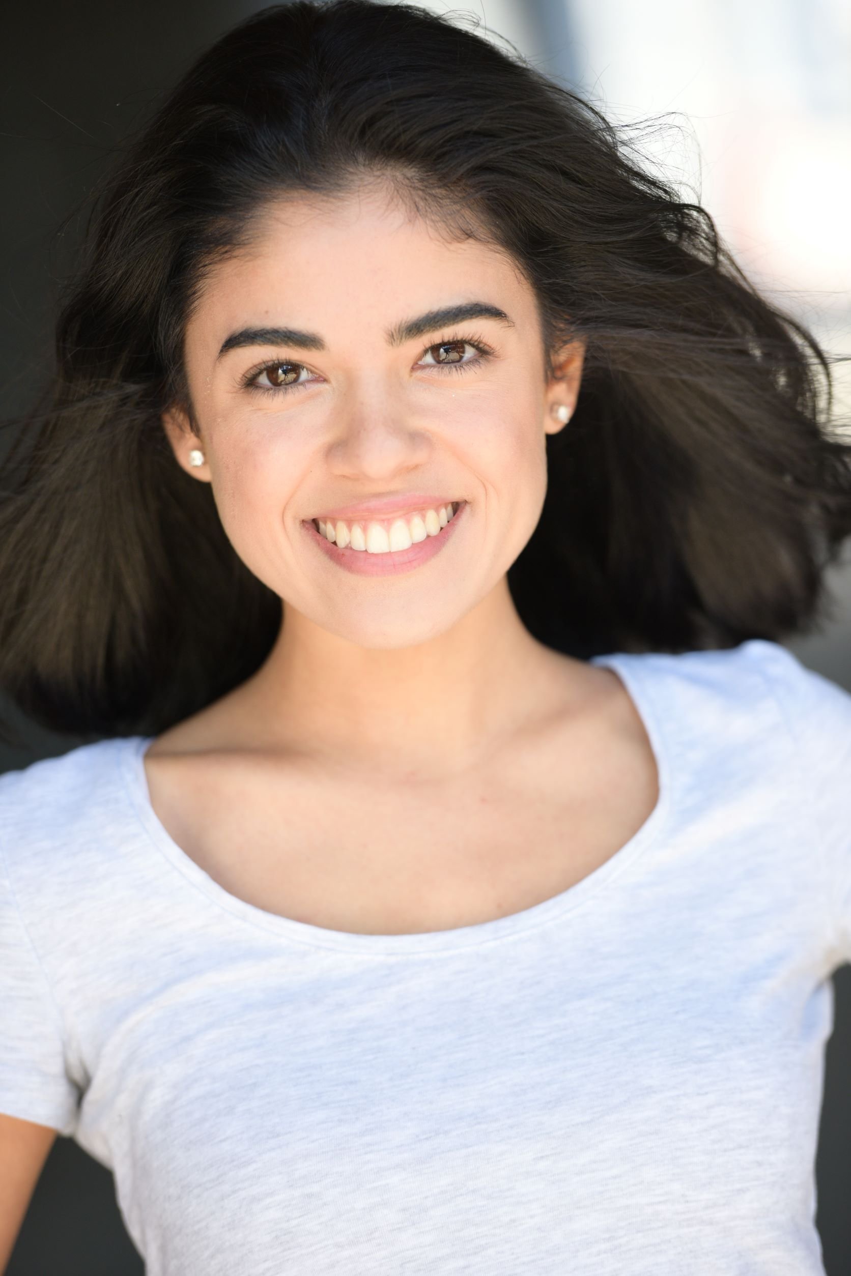 Young woman actress with dark hair smiling outdoors in a white T-shirt.