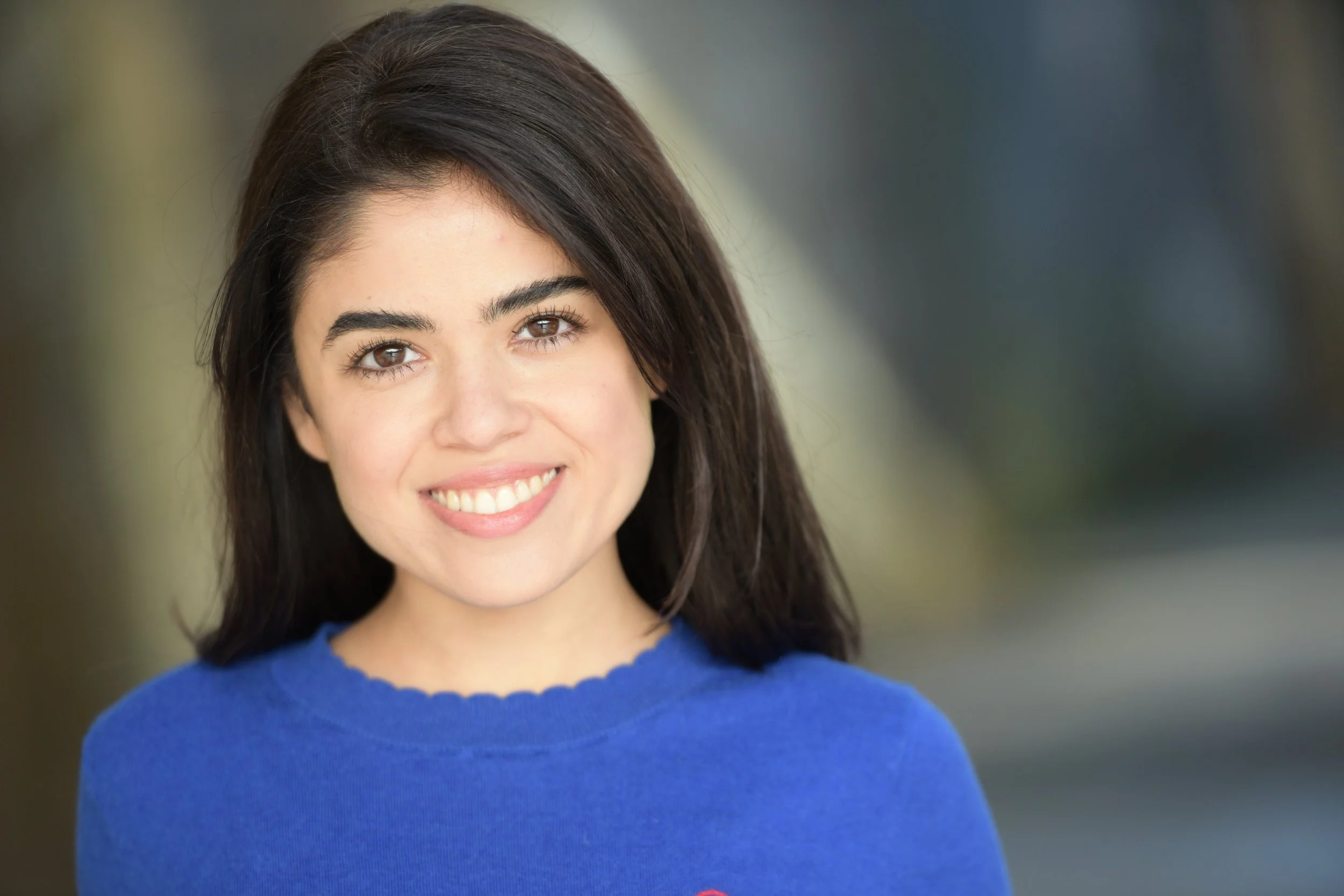 A woman actress with long dark hair smiling, wearing a blue top, outdoors with blurred background.