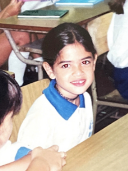 A young girl with dark hair and a blue and white school uniform sitting at a desk in a classroom.