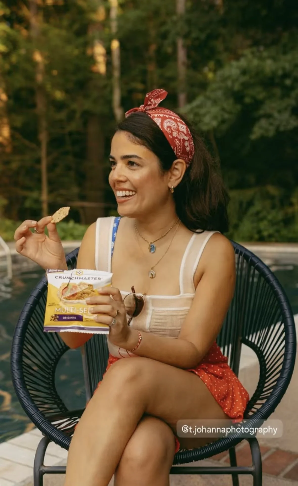 Petite female model with dark brown hair, brown eyes, and light skin, wearing a cute summer outfit featuring fourth of july look including a red bandana headband, a red zara polka dot skirt, and a shein cropped knit top. Eating multi-seed chips crisp