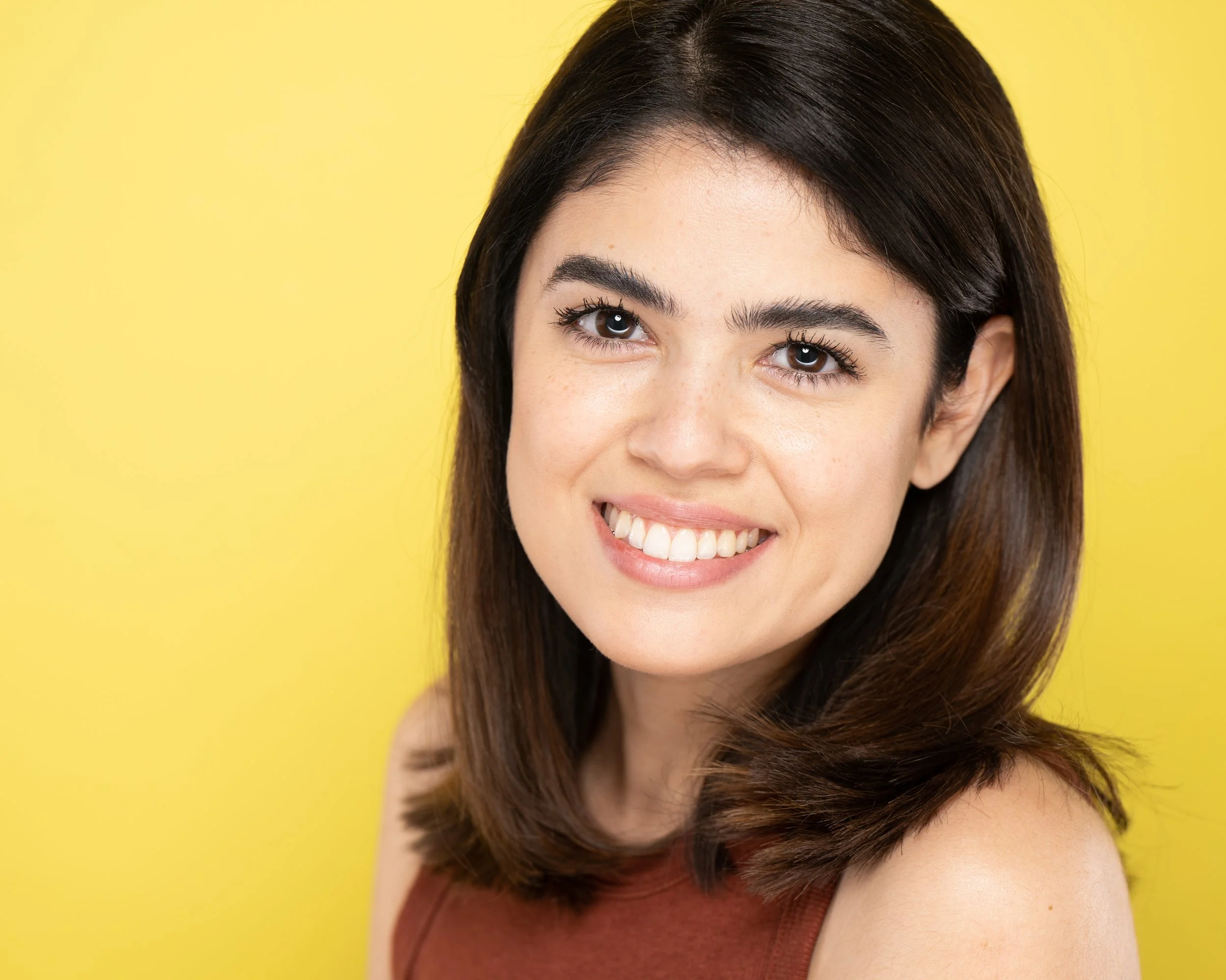 Close-up actor headshot of a smiling woman actress with shoulder-length brown hair, wearing a sleeveless rust-colored top, set against a yellow background.