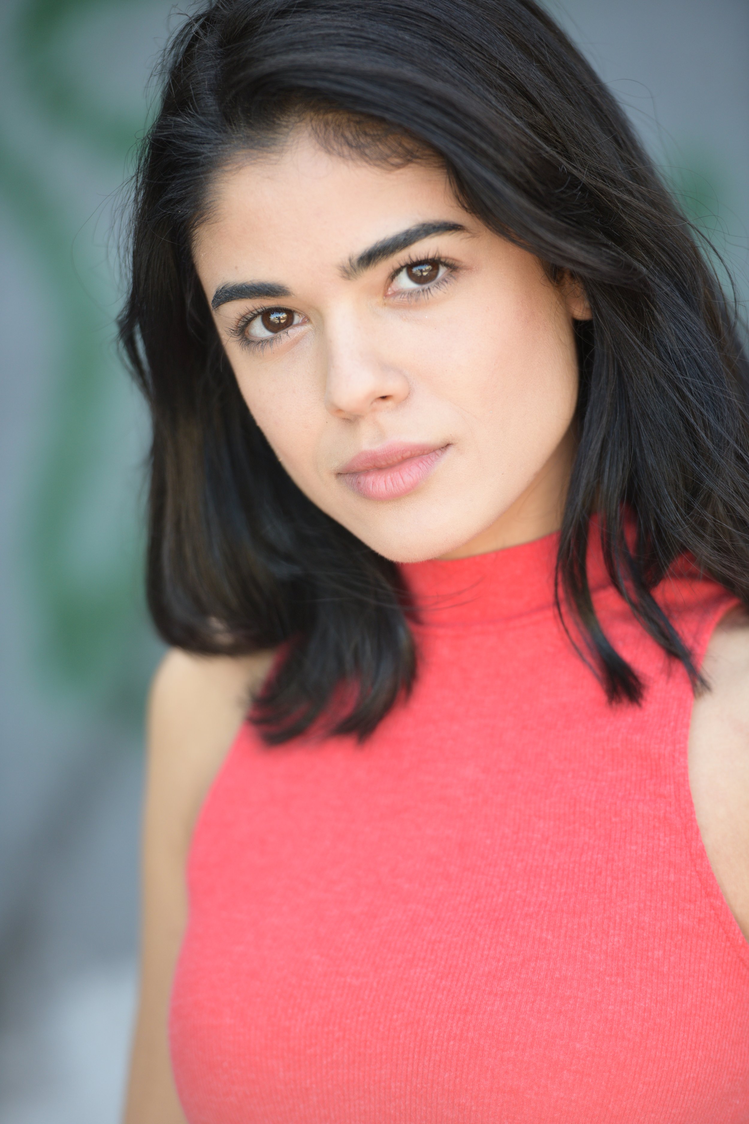 Close-up actor headshot of a young woman with dark hair, wearing a red sleeveless top, looking at the camera.