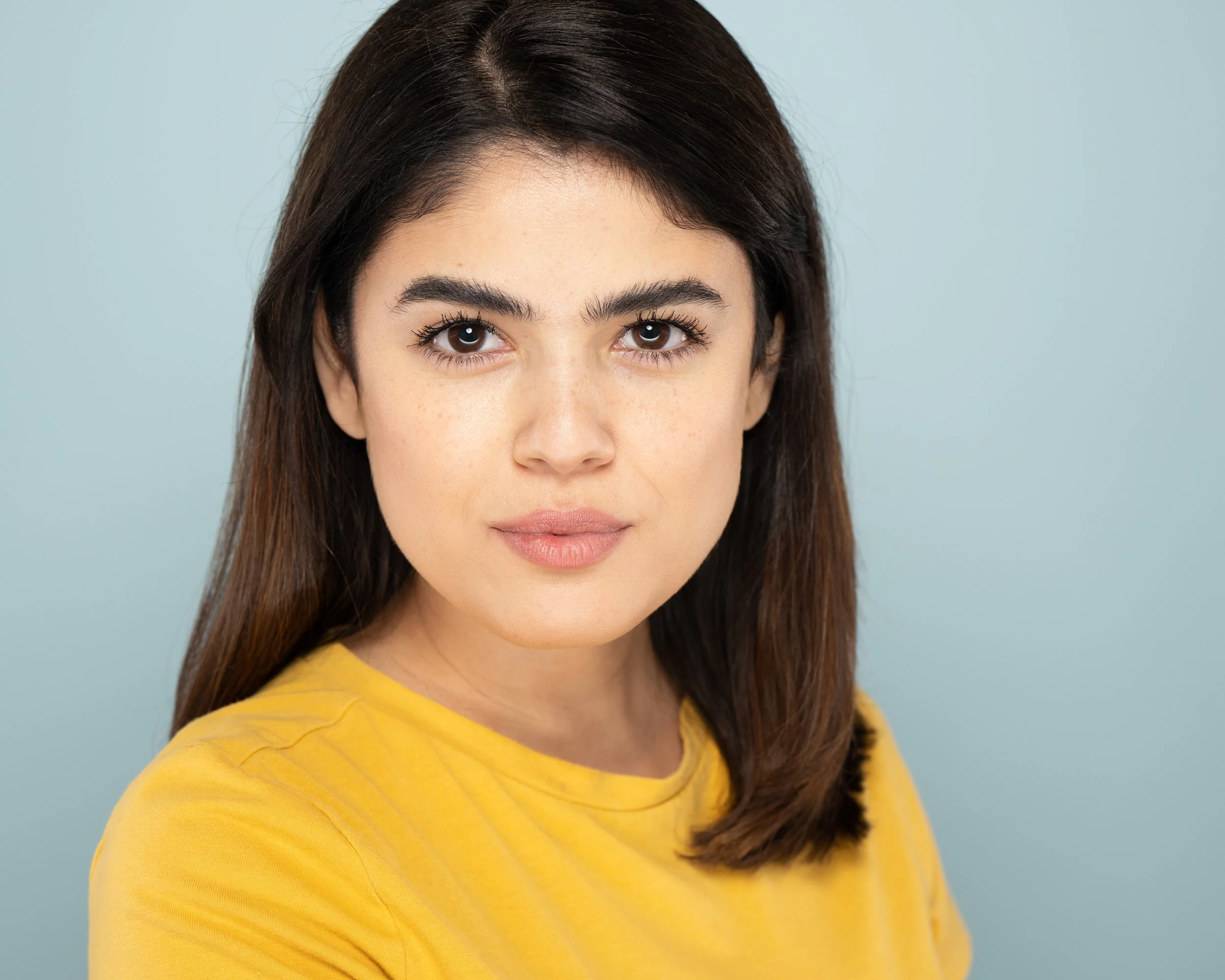 A woman actress with dark brown hair, wearing a yellow shirt, against a light blue background.