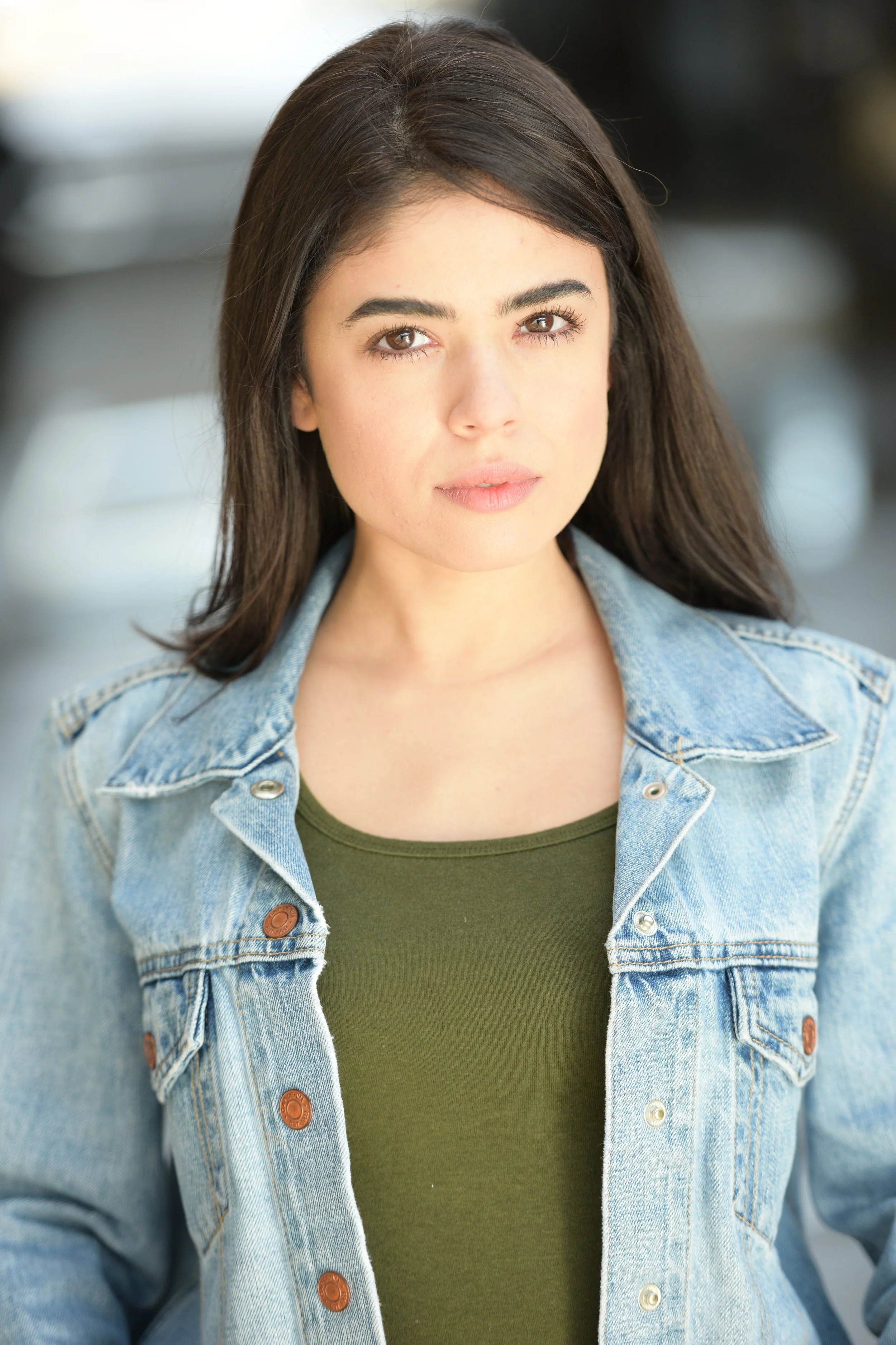 A young woman actress with long dark hair, wearing a denim jacket over a green top, looking directly at the camera with a neutral expression.