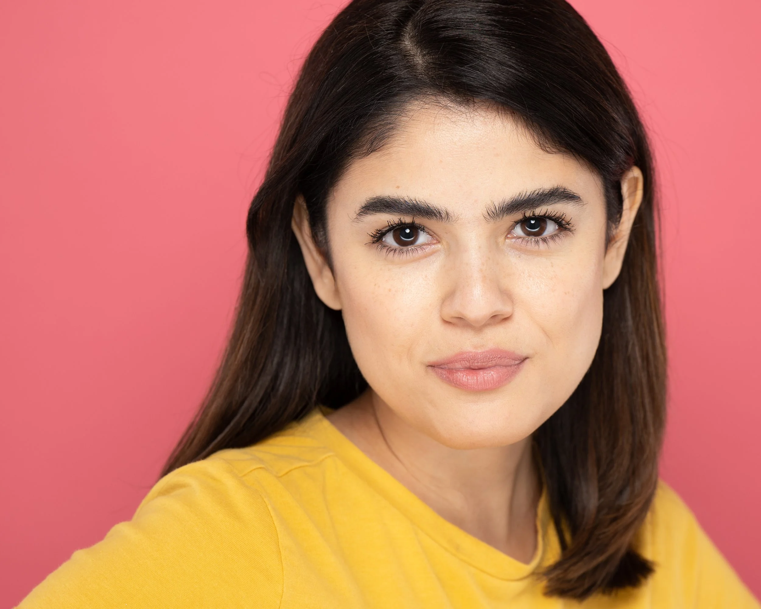 A young woman actress with brown hair, wearing a yellow shirt, standing against a pink background.