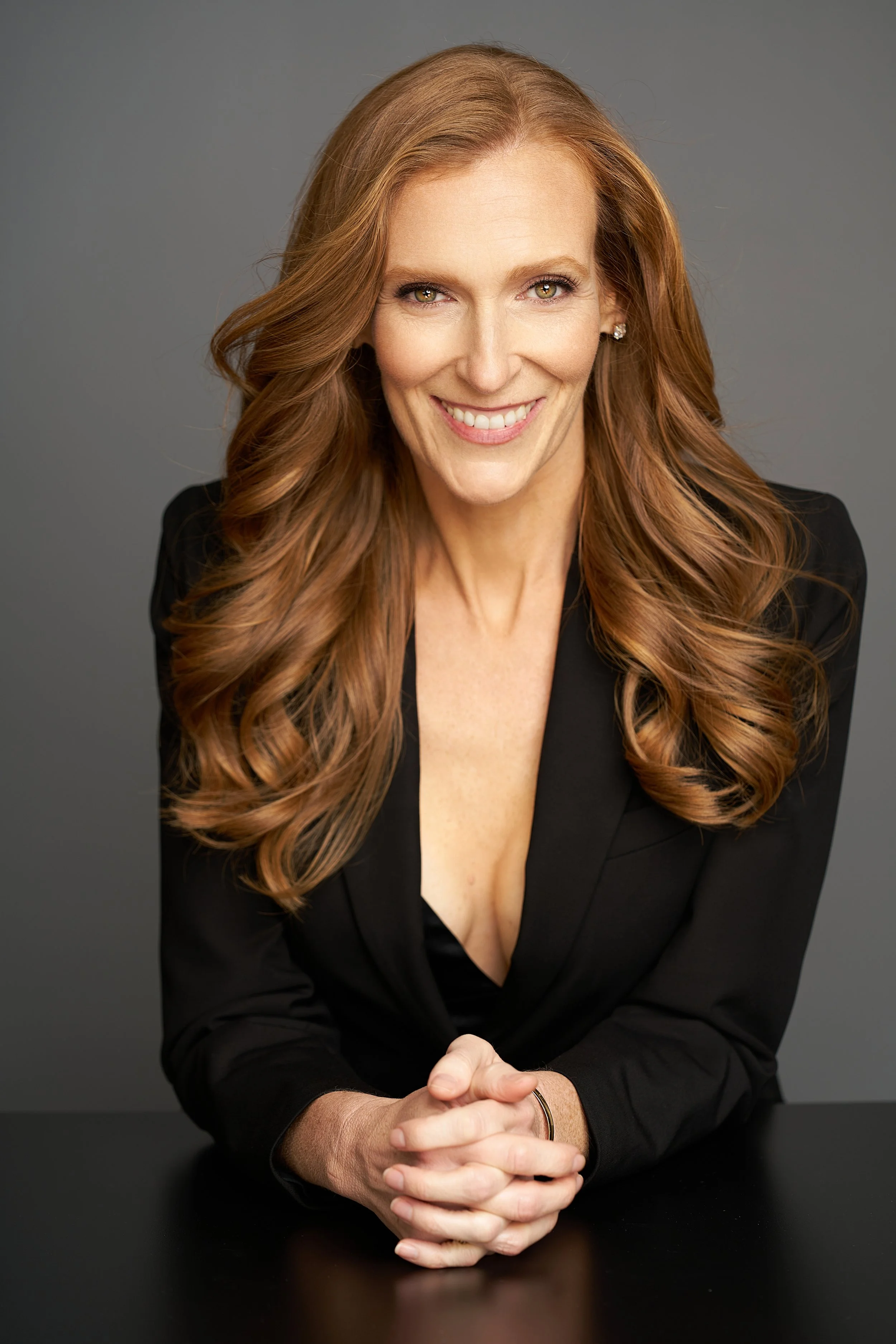 Portrait of a woman with long, wavy red hair smiling and sitting at a black table against a gray background.