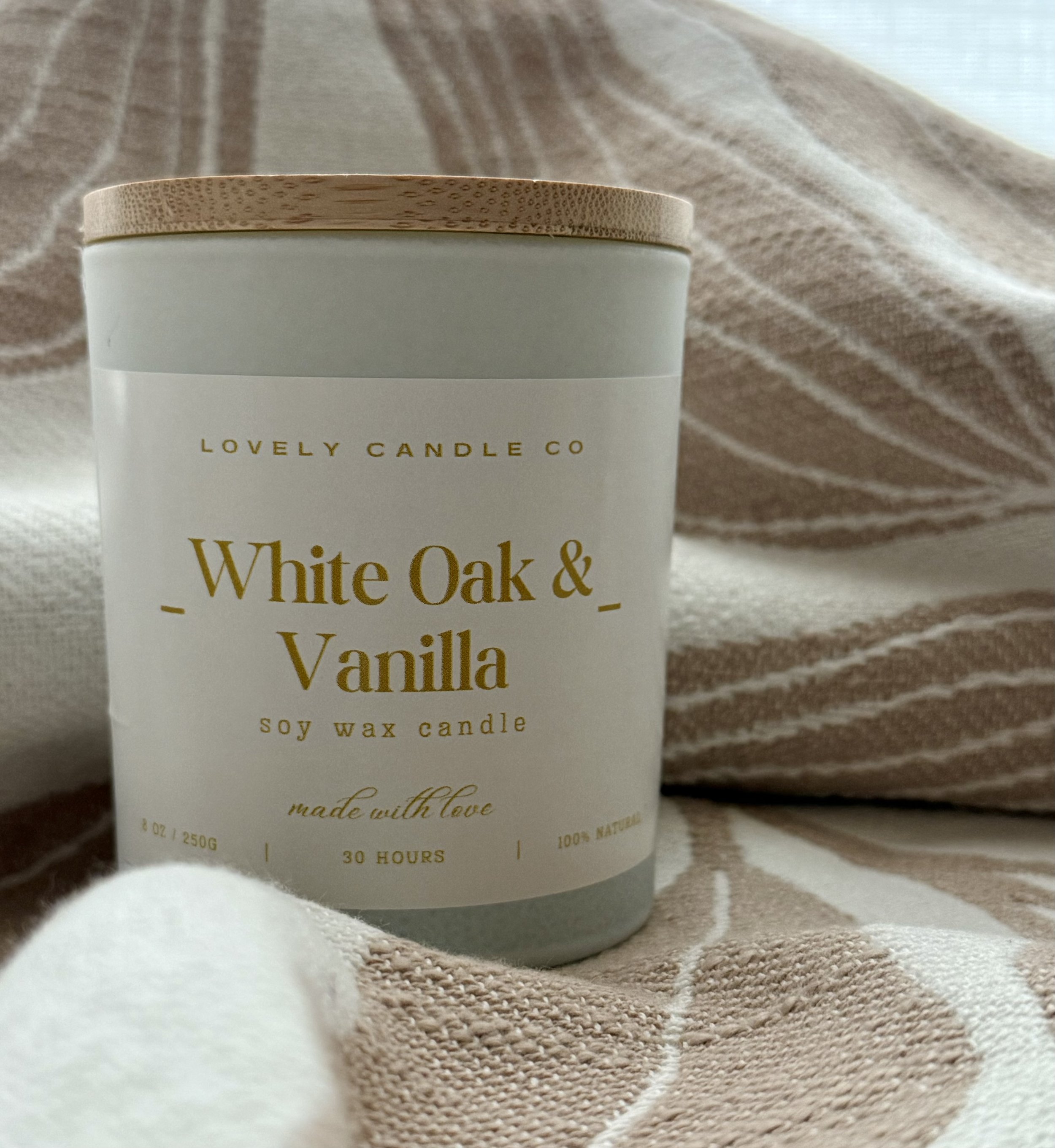 A white soy wax candle labeled 'White Oak & Vanilla' from Lovely Candle Co, placed on a textured beige and white striped fabric background.