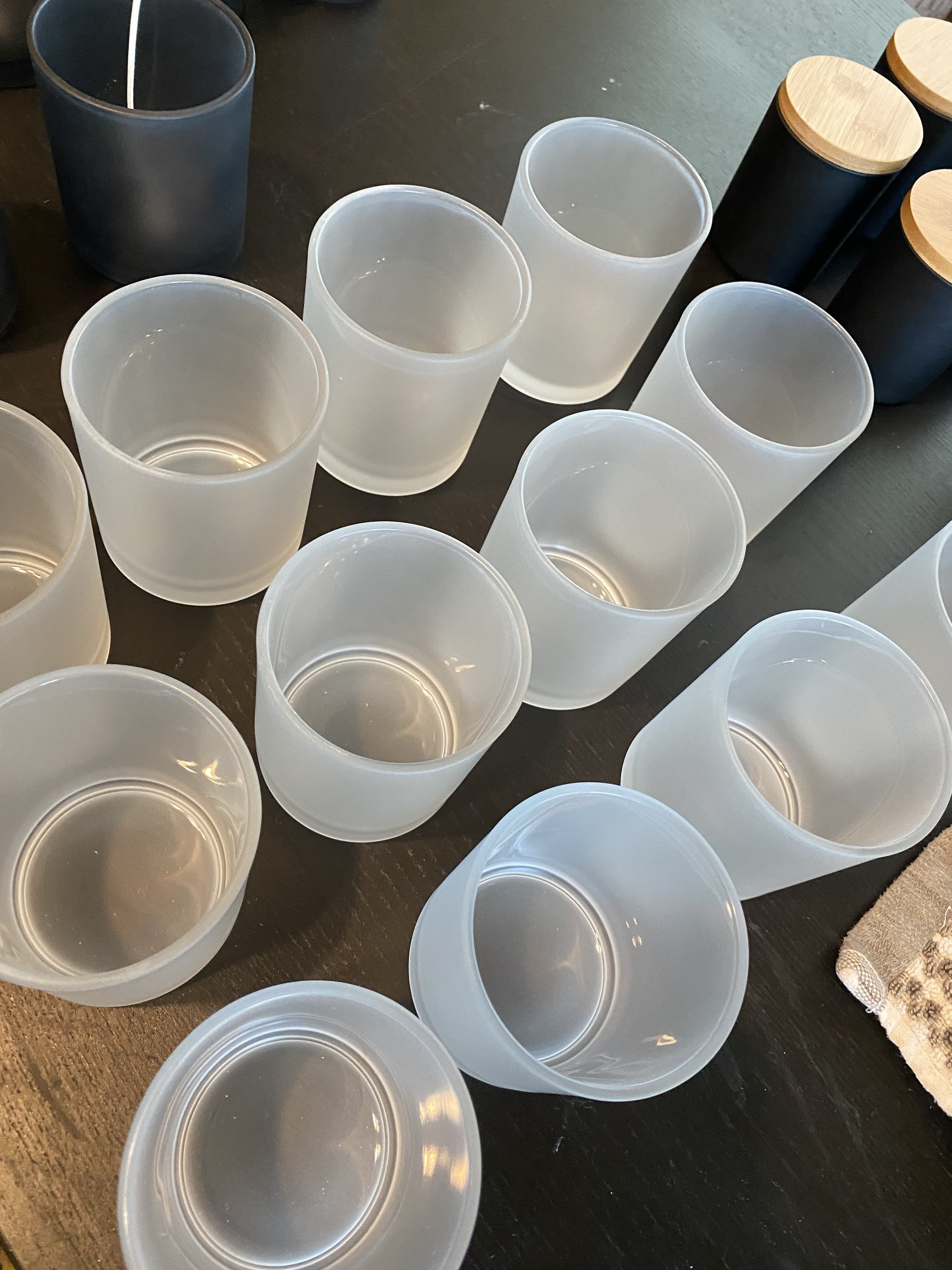 Multiple frosted glass candle vessels arranged on a black table, with some white candle vessels and black candle vessels with wooden lids in the background.