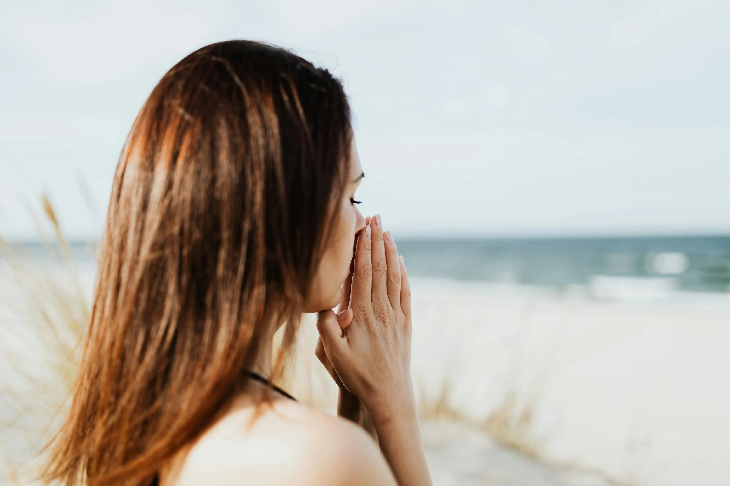 Woman praying on a beach by the ocean with her eyes closed and hands clasped