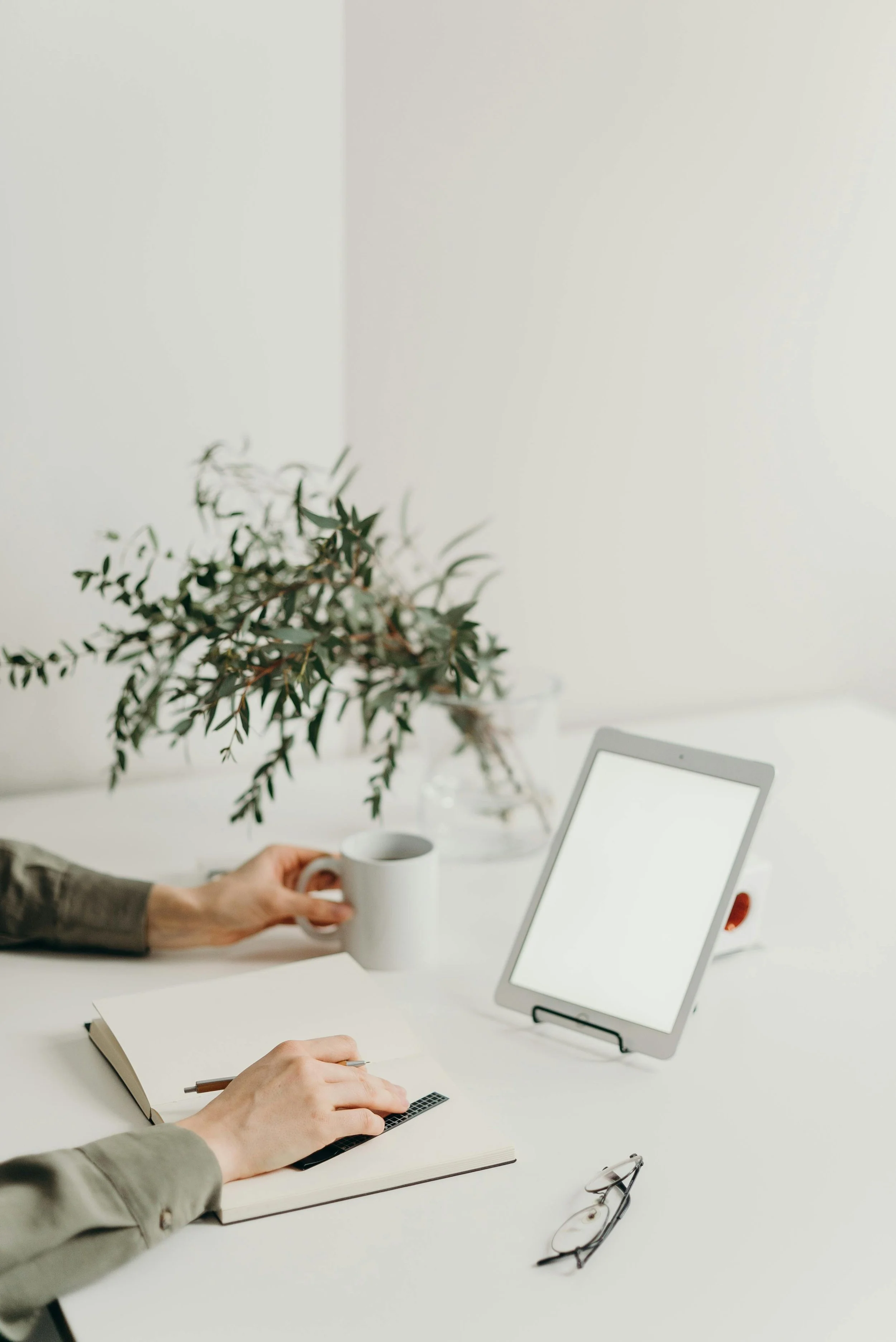 Person writing in a notebook with a pencil, sitting at a white desk, holding a mug, with a tablet, glasses, and a plant in the background.