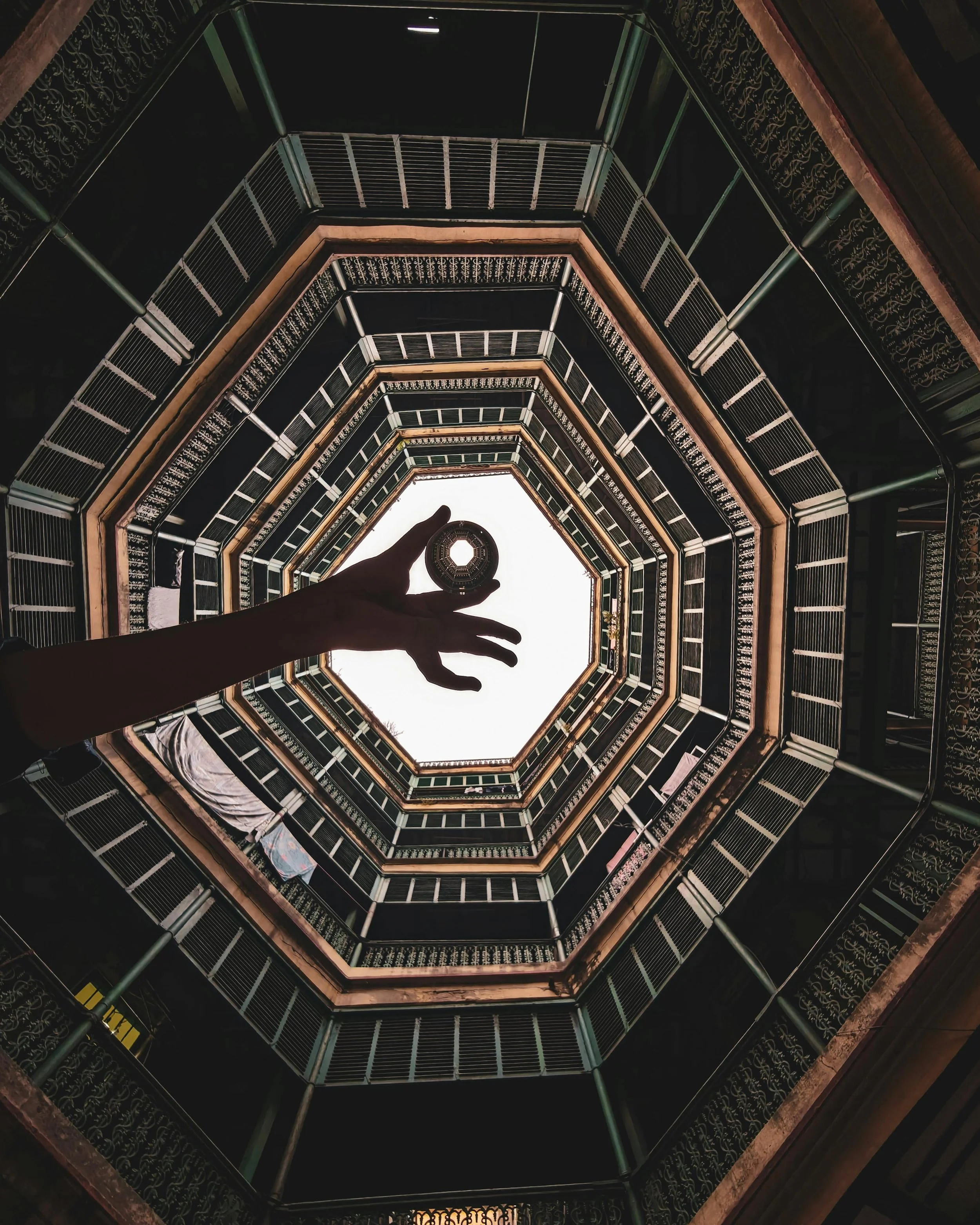 A person holding a coin in the center of the camera, with the view looking up through an open, multi-story stairwell to the sky.