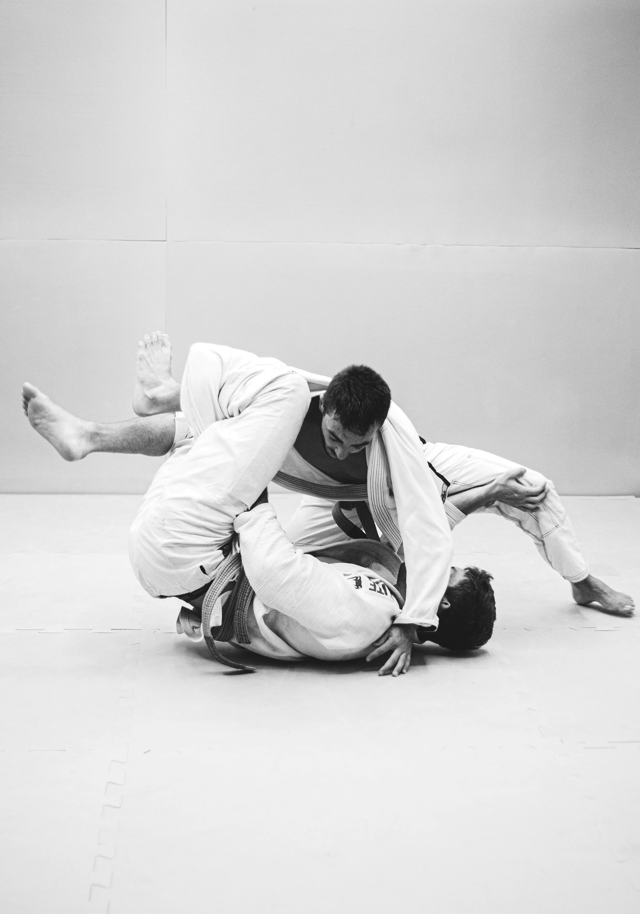 Two men in jiu-jitsu gis grappling on a mat, with one man in the process of executing a submission move on the other.