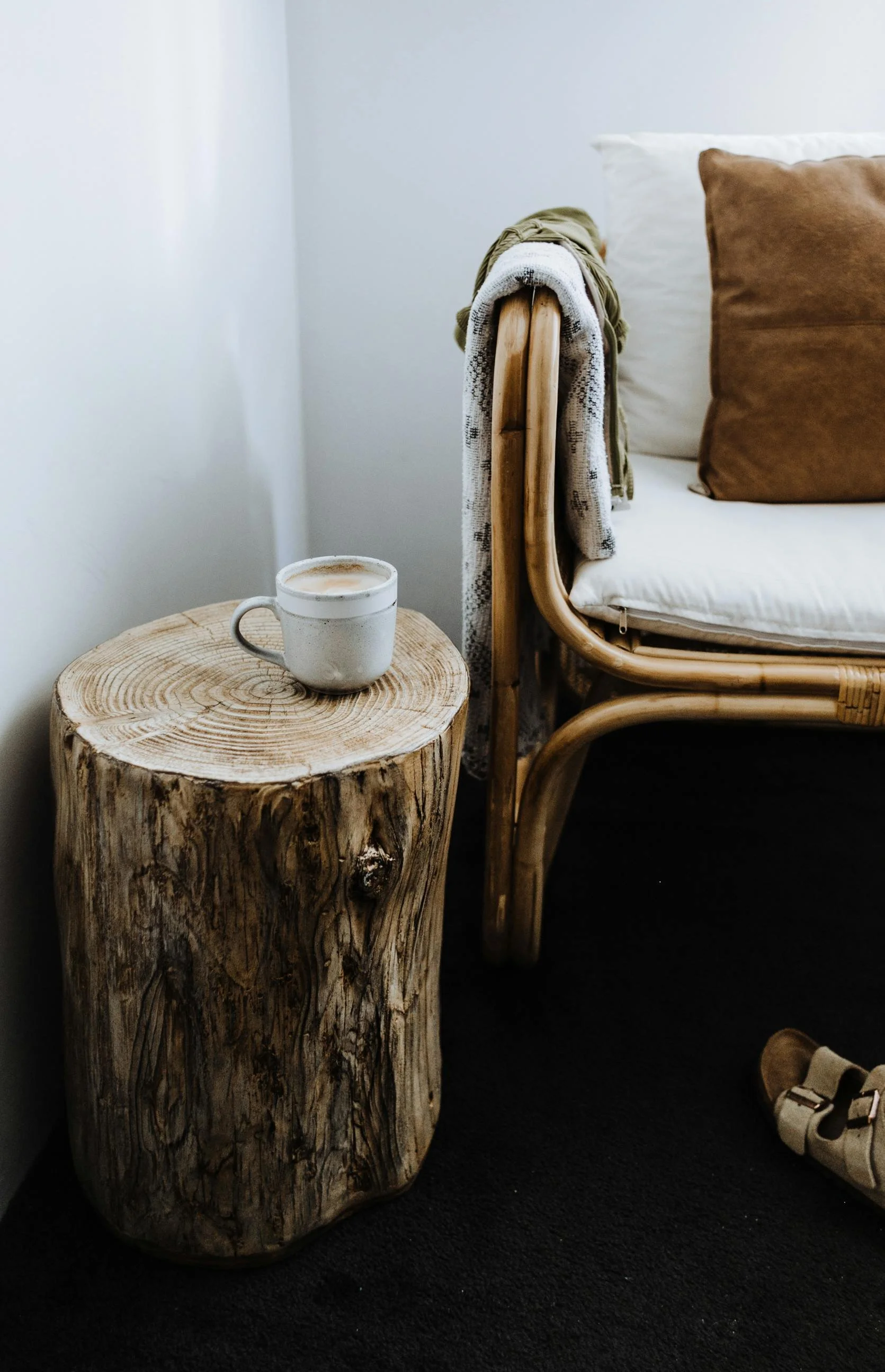 Close-up of a bedside table made from a large, natural wood log with a coffee mug on top, next to a rattan bed frame with beige and brown pillows.