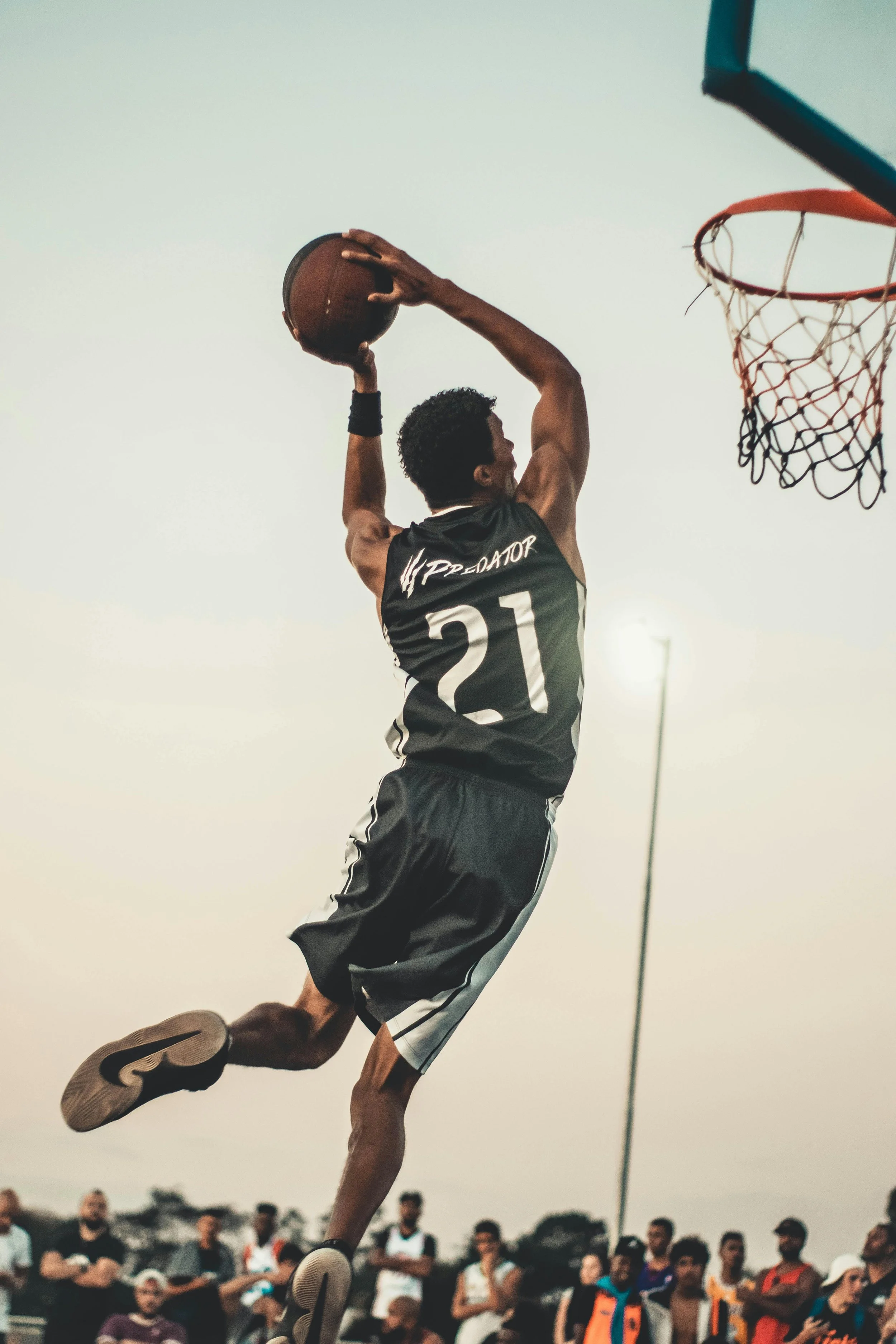 A young man wearing a black sports jersey with the number 21 is mid-air, dunking a basketball towards the hoop during an outdoor game, with spectators watching in the background.