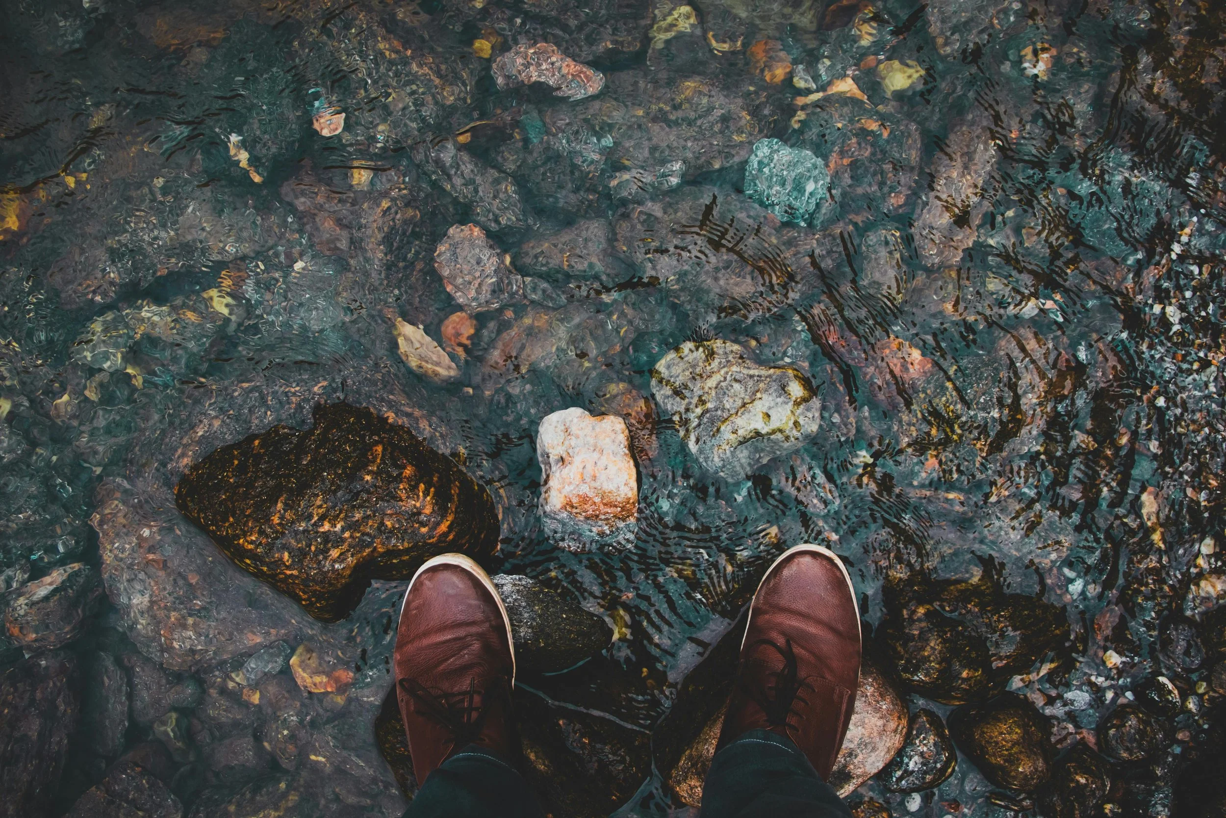 Person standing on rocks at the edge of a water body with visible ripples and submerged stones.