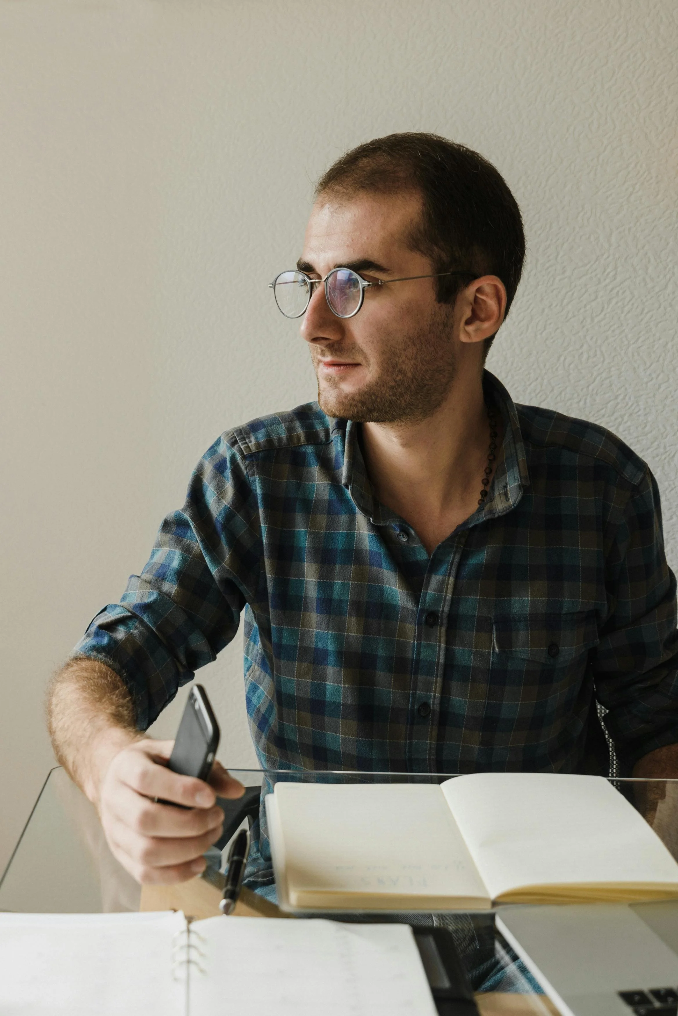A young man with glasses and a plaid shirt sitting at a desk with an open notebook, a pen, a smartphone in his hand, and a closed laptop.
