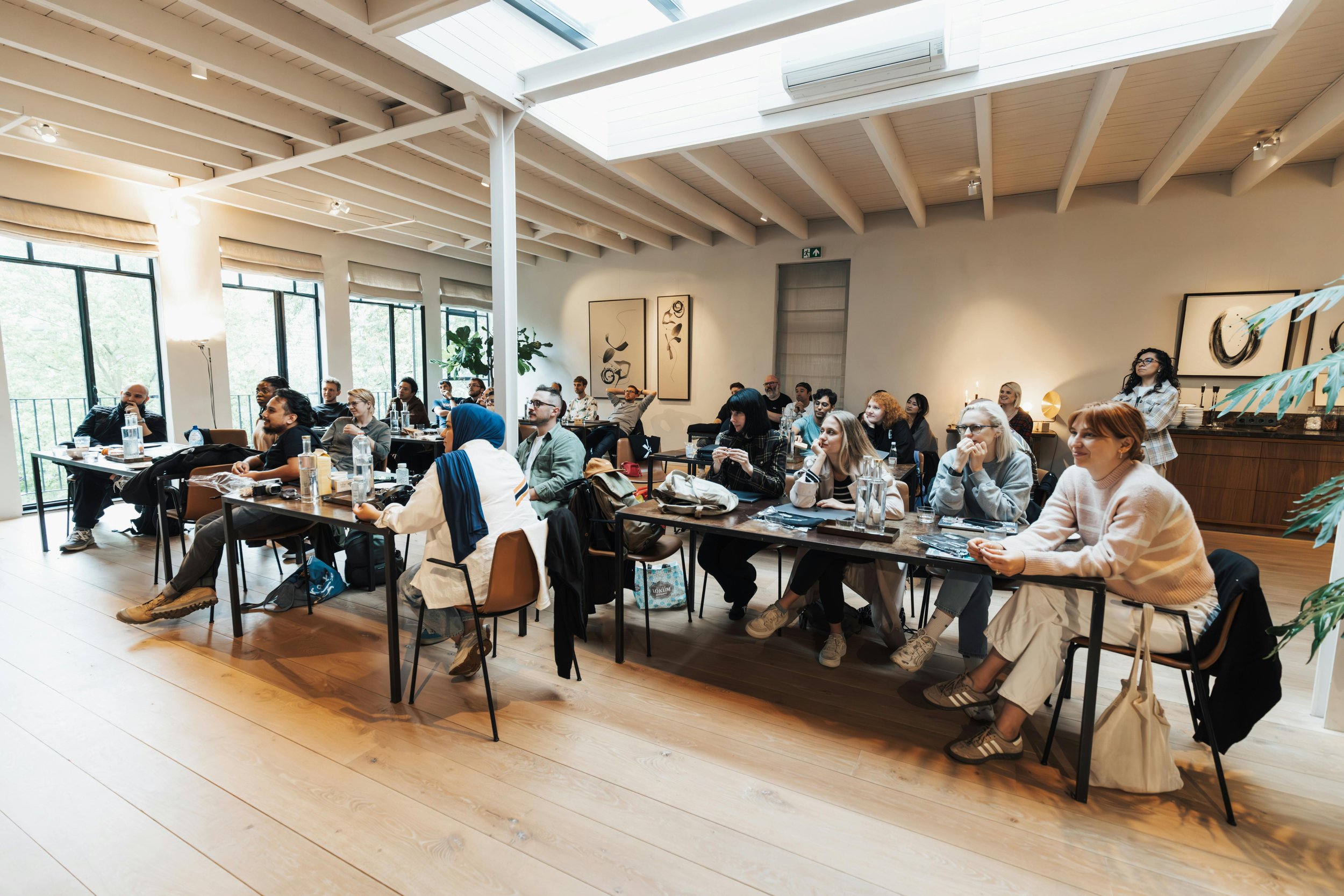 A group of diverse people attending a conference or workshop in a well-lit modern room with large windows, seating at tables with water bottles, notebooks, and snacks.