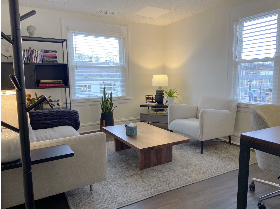 A cozy living room with two white armchairs and a wooden coffee table, a side table with a lamp and plants, and a bookcase with books on the left. There are two windows with blinds allowing natural light to brighten the room.