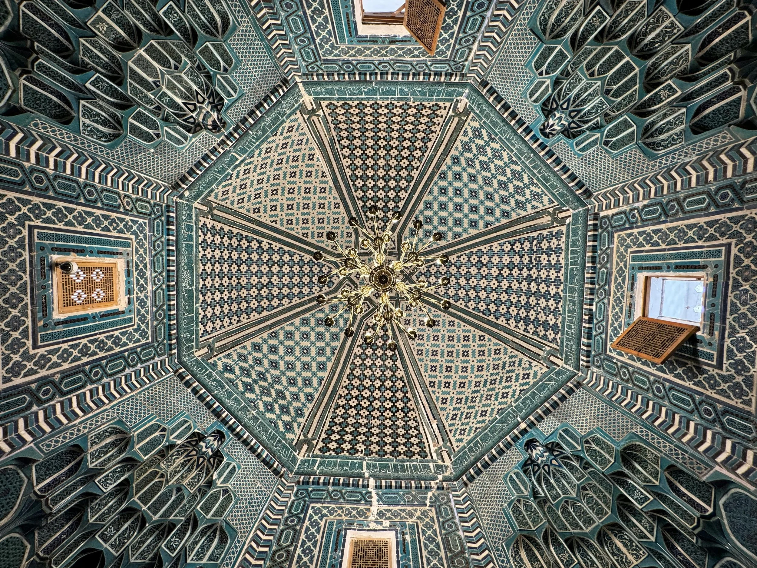 Intricate ornately decorated ceiling with geometric blue and white tile patterns, a central chandelier, and two windows with wooden lattice covers.