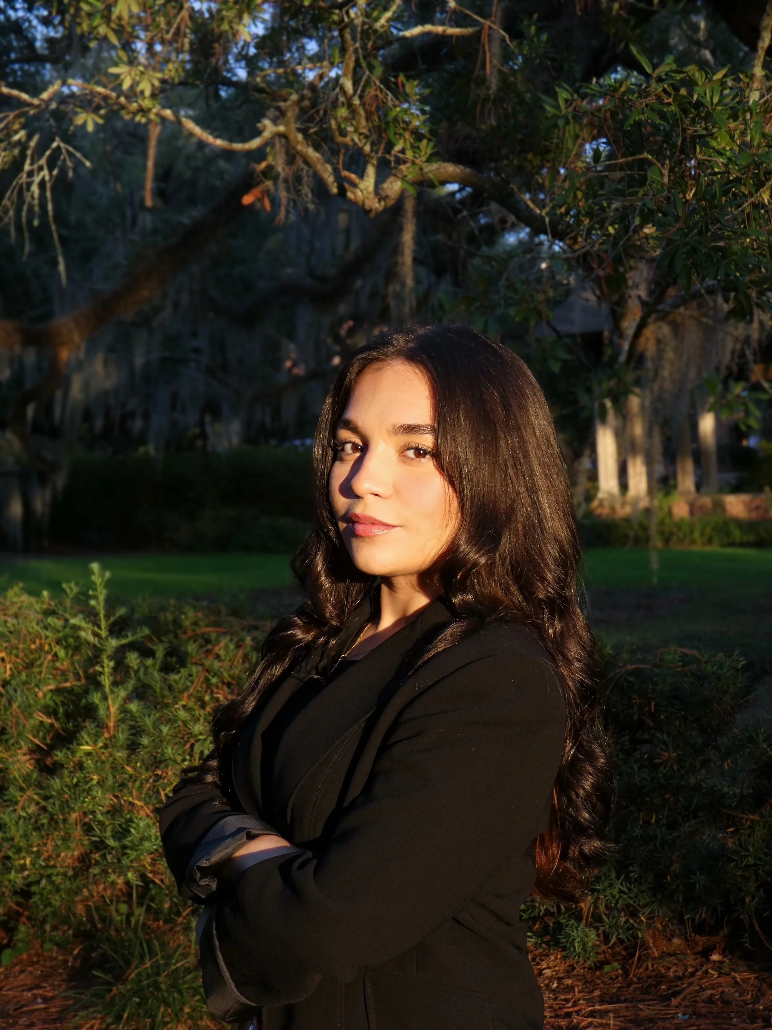 A woman with long dark hair standing outdoors in sunlight, with a background of trees and bushes.