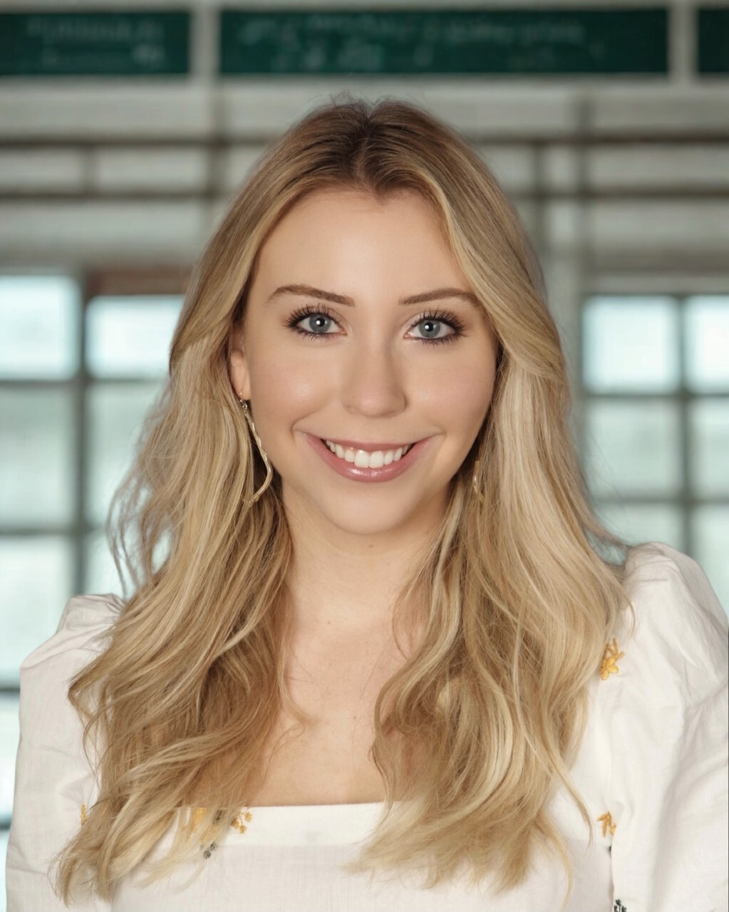 A woman with long blonde hair and blue eyes smiling indoors with windows in the background.