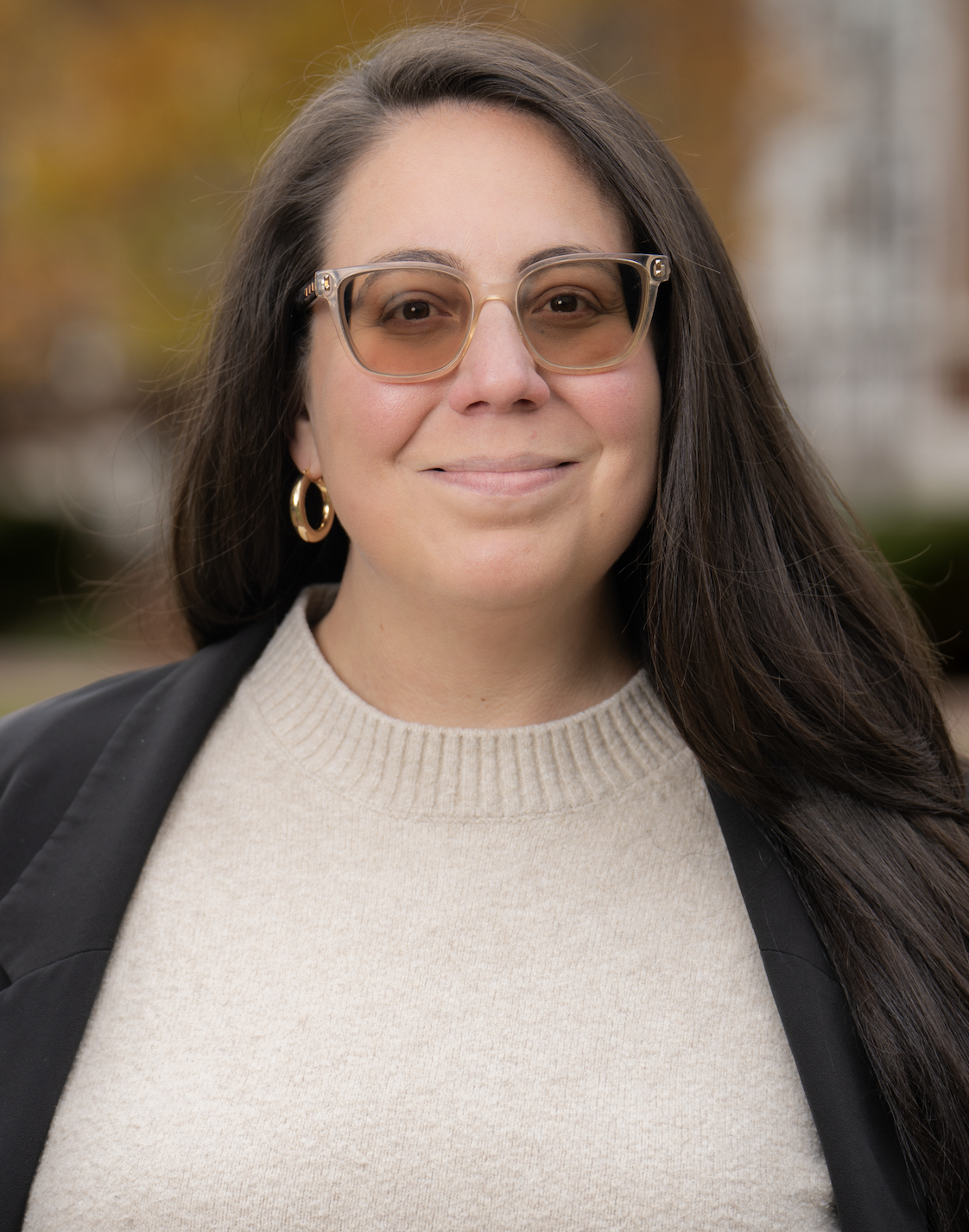 A woman with long dark hair, wearing sunglasses, gold hoop earrings, a beige sweater, and a black jacket outdoors during fall.