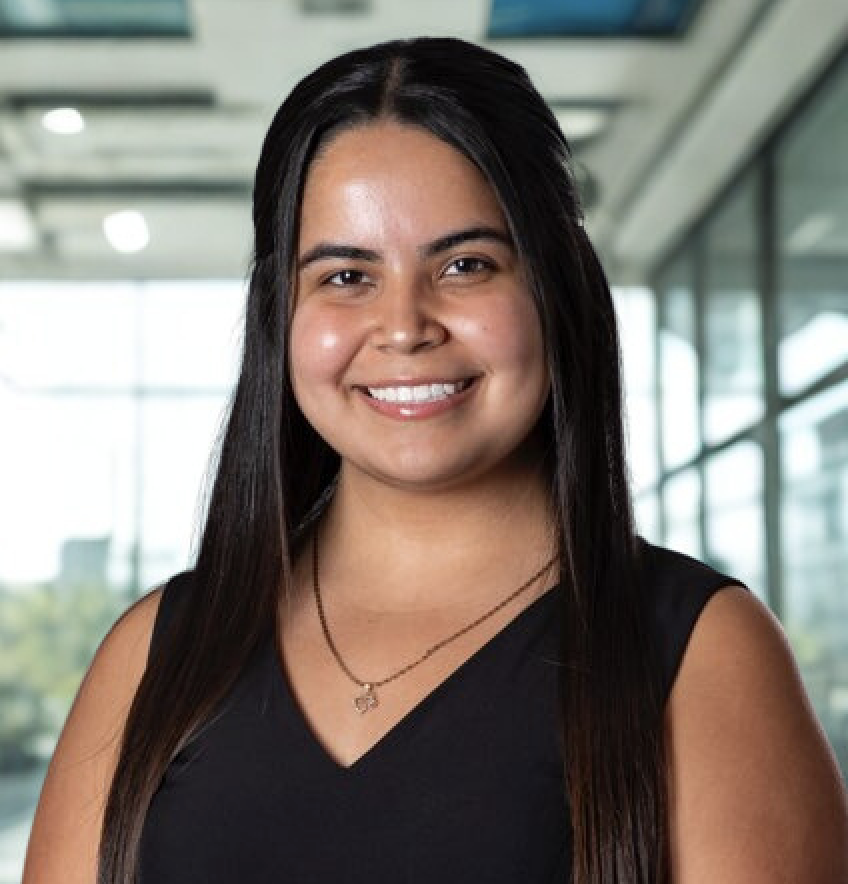 A smiling young woman with long black hair wearing a black sleeveless top and a necklace, standing indoors with large windows and a modern building interior in the background.