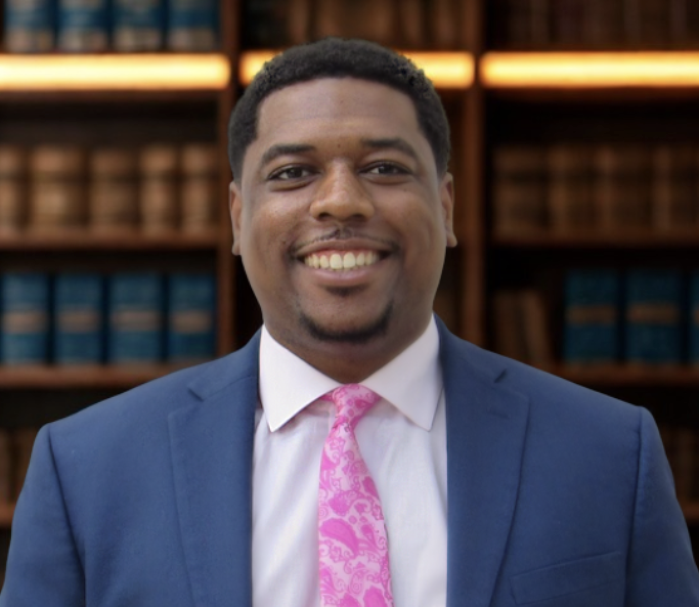 A smiling man in a blue suit, white shirt, and pink tie standing in front of a bookshelf filled with books.