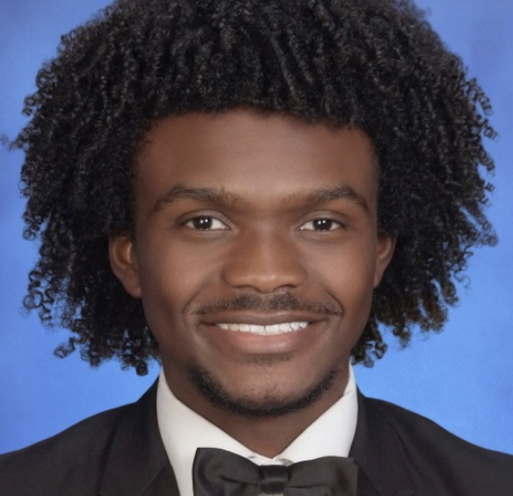 A young man with dark curly hair, smiling, wearing a tuxedo with a bow tie, posed for a professional portrait against a blue background.