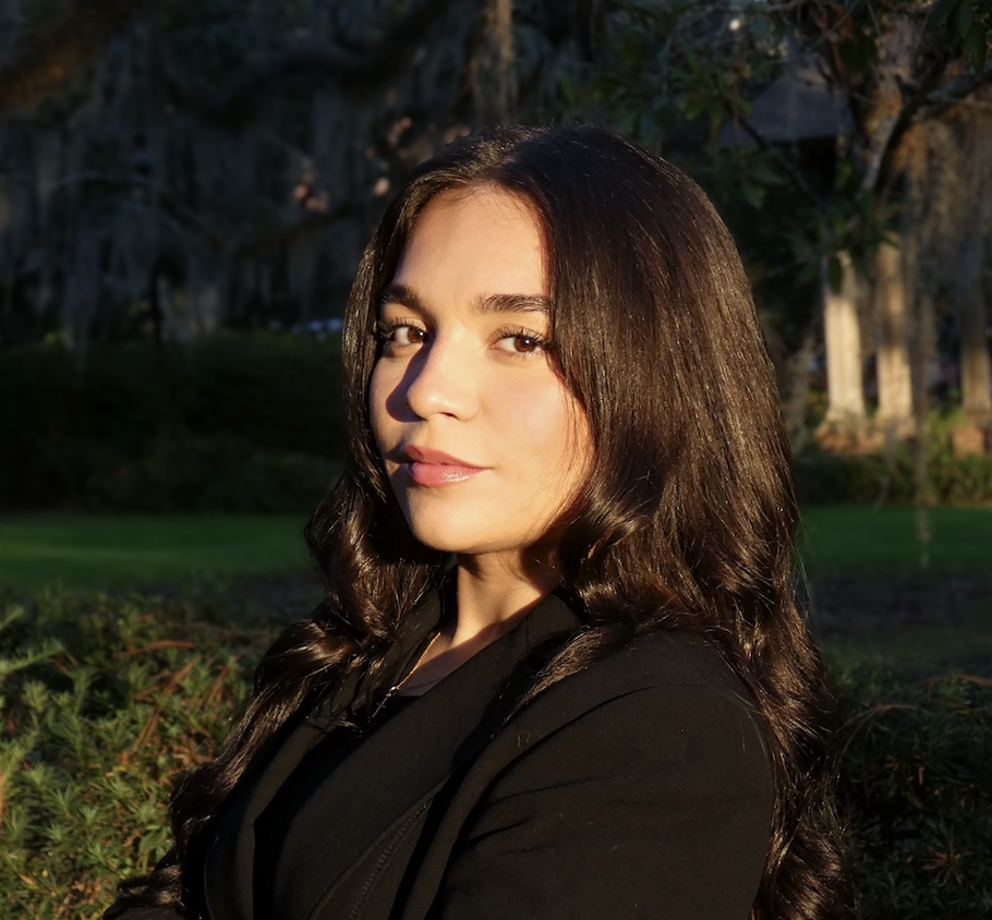 A woman with long, wavy dark hair, wearing a black top, stands outdoors during sunset with trees and greenery in the background.