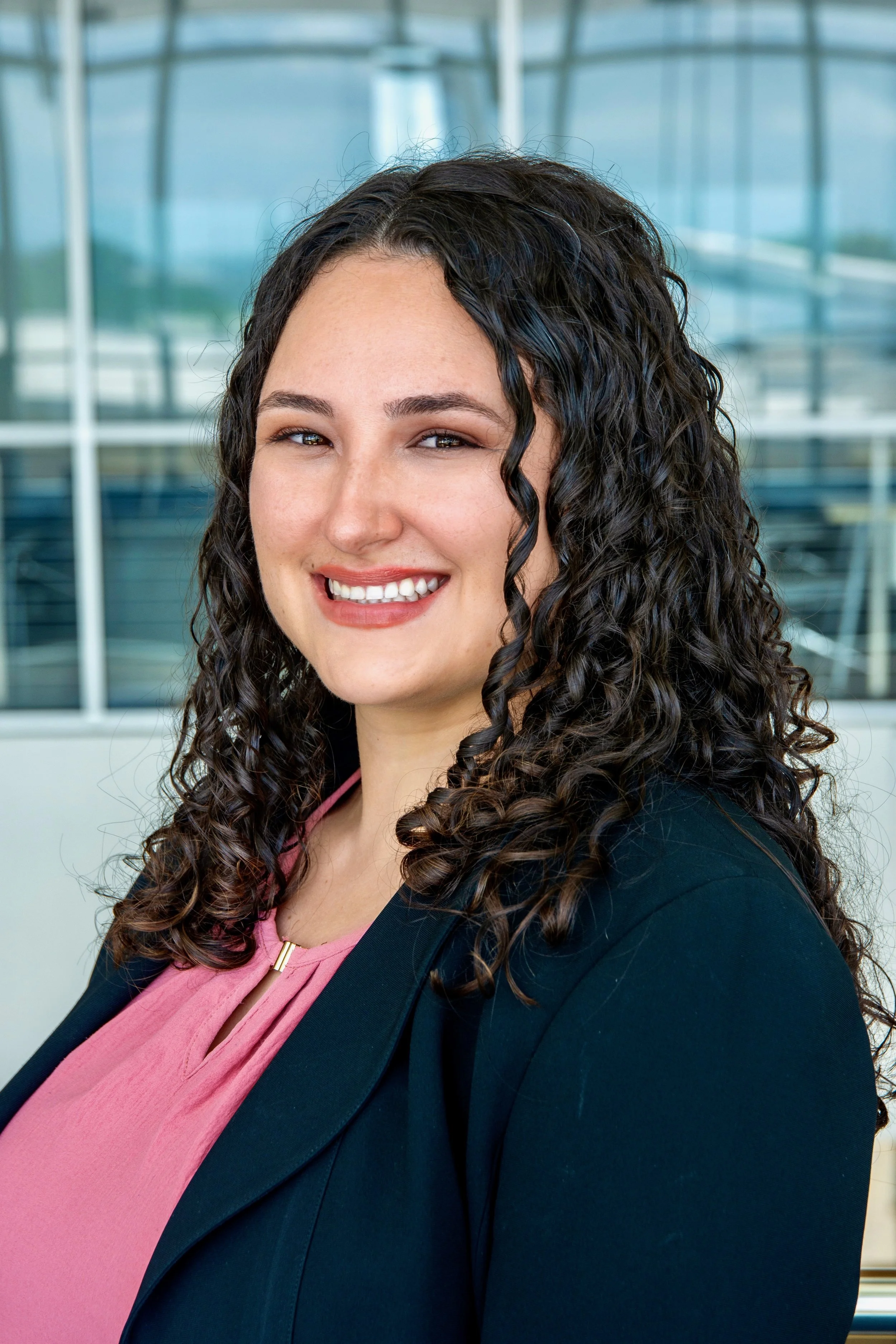 A woman with long, dark, curly hair smiling at the camera inside a modern building with glass windows.
