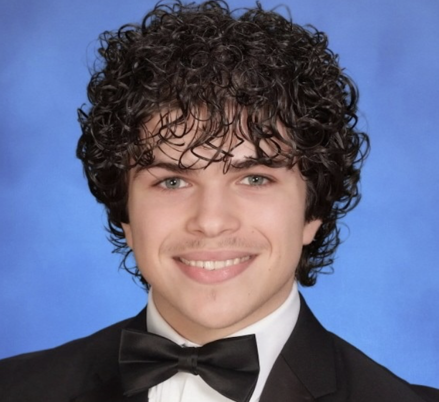 A young man with curly dark hair, light skin, and blue eyes wearing a tuxedo and black bow tie, smiling against a blue background.