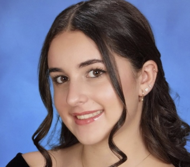 Close-up portrait of a young woman with dark brown hair styled in loose curls, smiling against a blue background.
