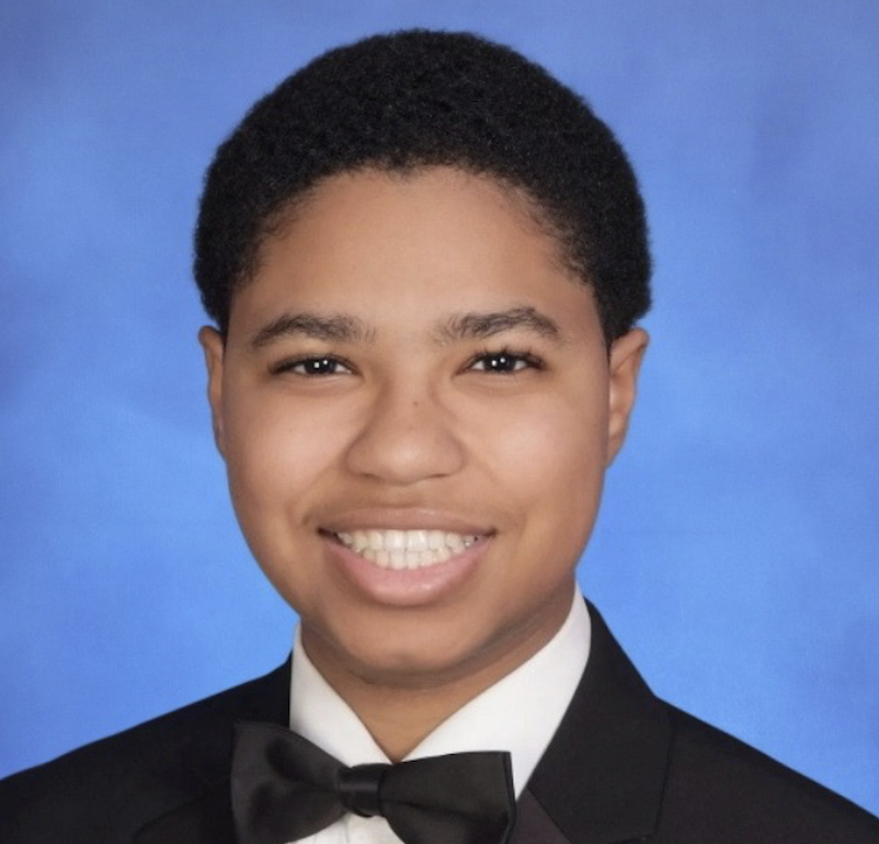 Young man with short curly hair wearing a tuxedo with a bow tie, smiling against a blue background.