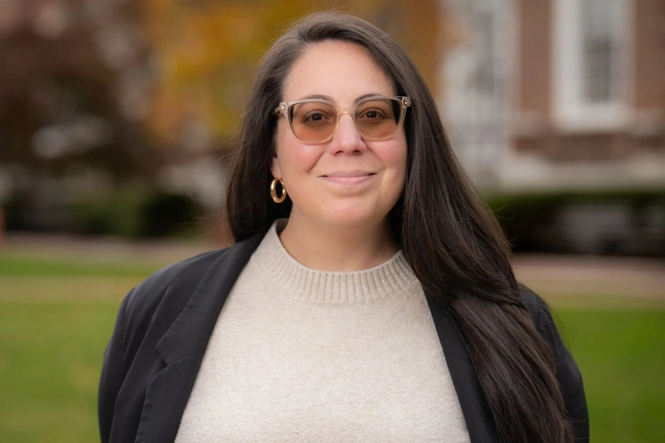 A woman with long dark hair, wearing glasses, gold hoop earrings, a beige sweater, and a black jacket, smiling outdoors with a park and buildings in the background.