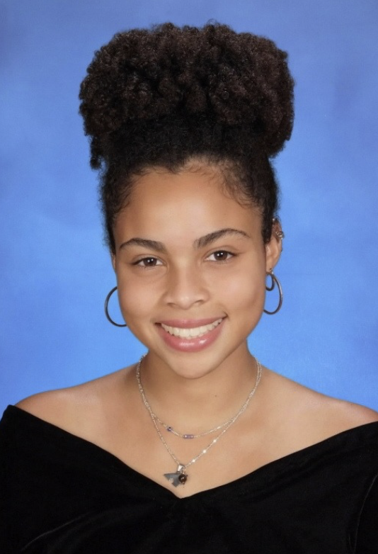 Young woman with a natural curly updo hairstyle, smiling, wearing hoop earrings and layered necklaces, against a blue background.